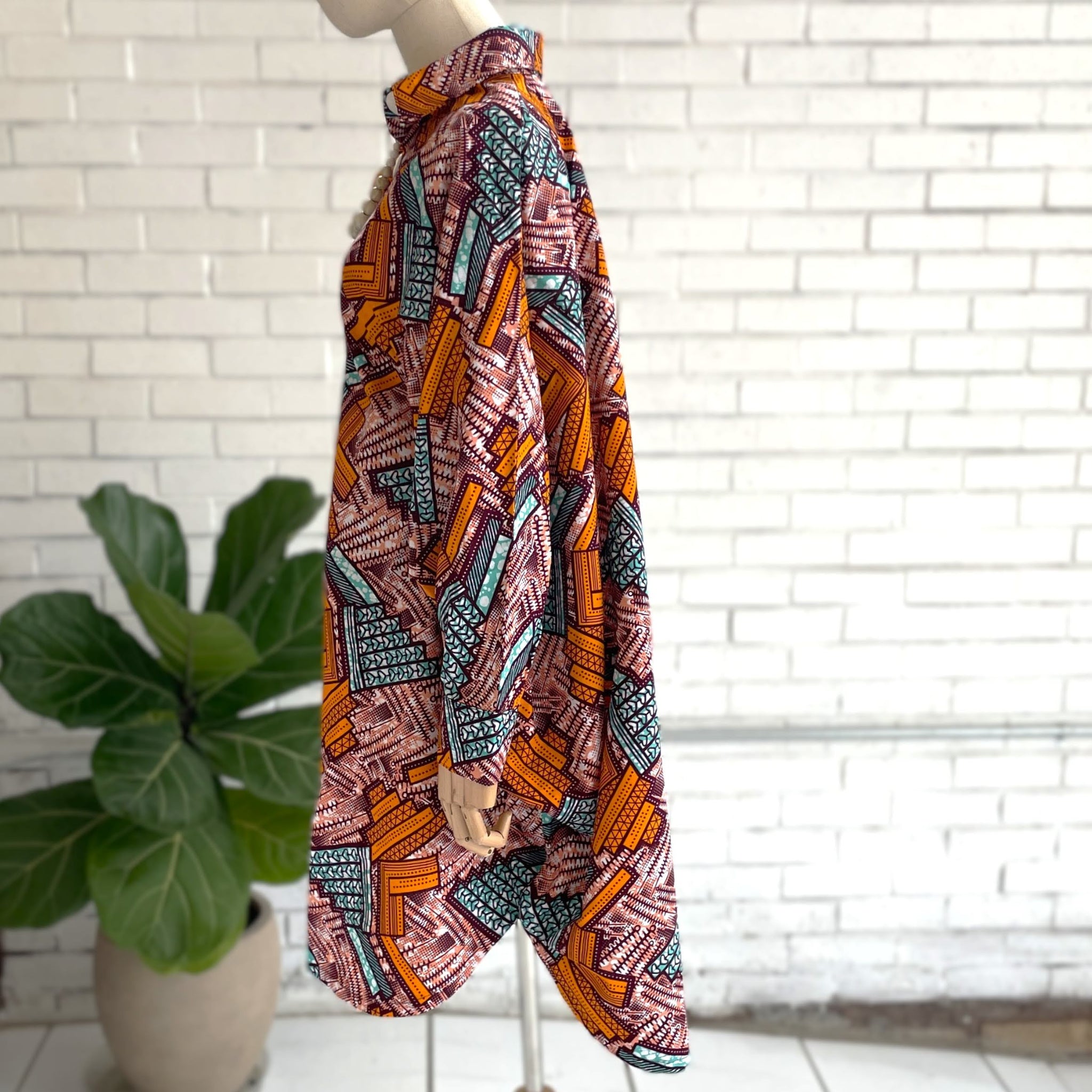 Colorful patterned dress on a mannequin against a white brick wall with a plant in the foreground.