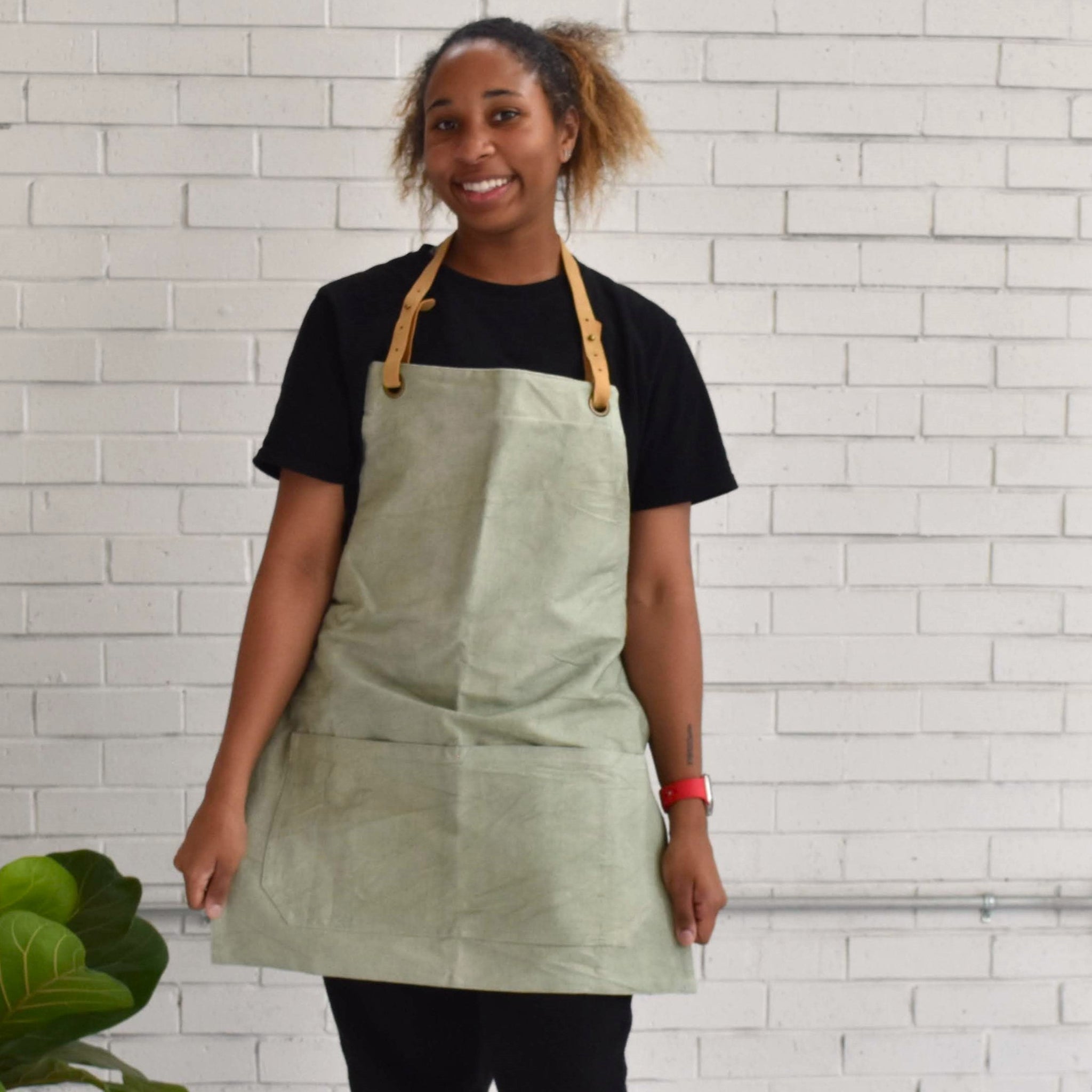 A woman wearing a sage green canvas apron with tan leather straps over a black t-shirt, standing in front of a white brick wall.