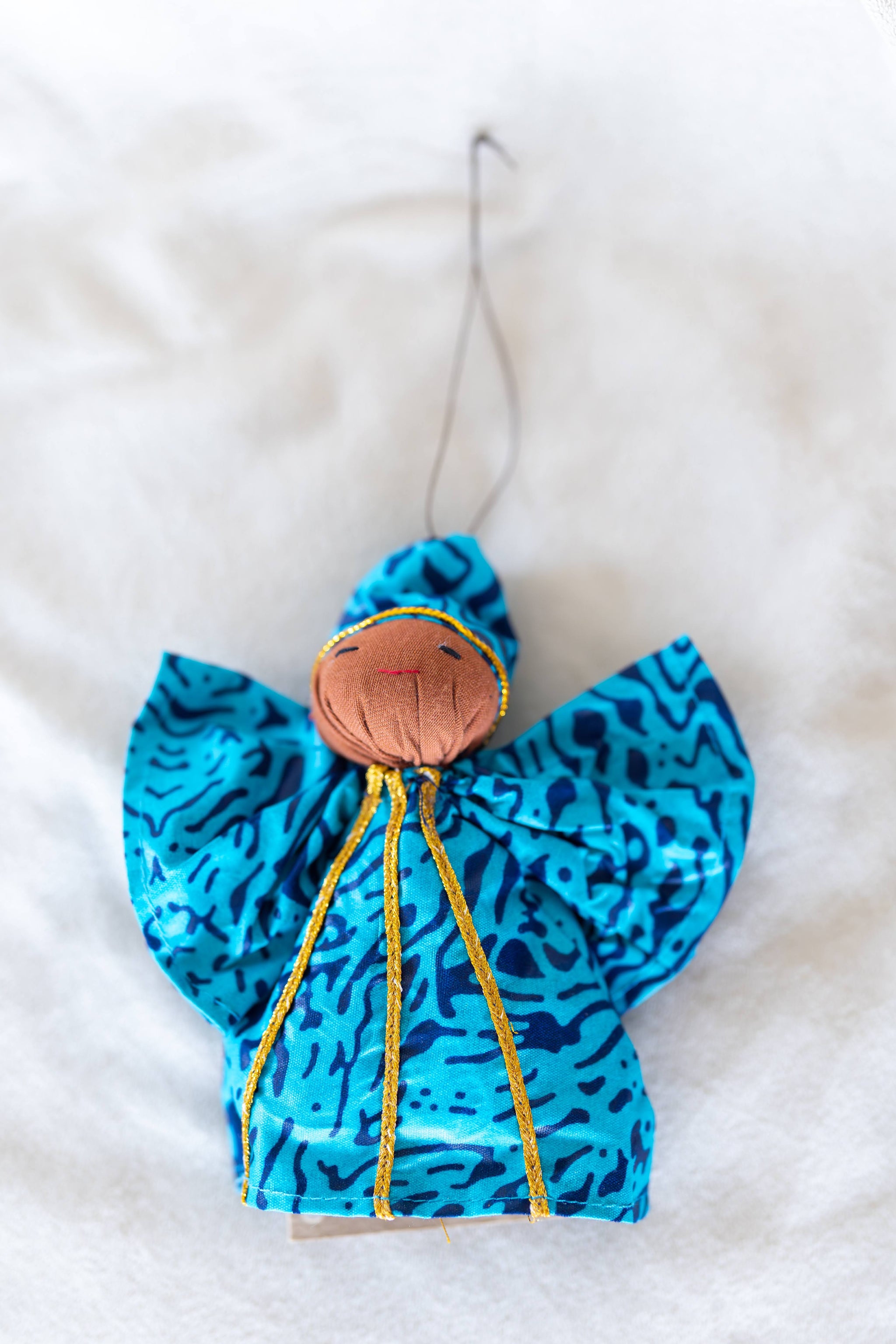 A handmade fabric angel ornament with bright blue and black patterned wings, a brown fabric head, and gold embroidered details, hanging against a white textured background.