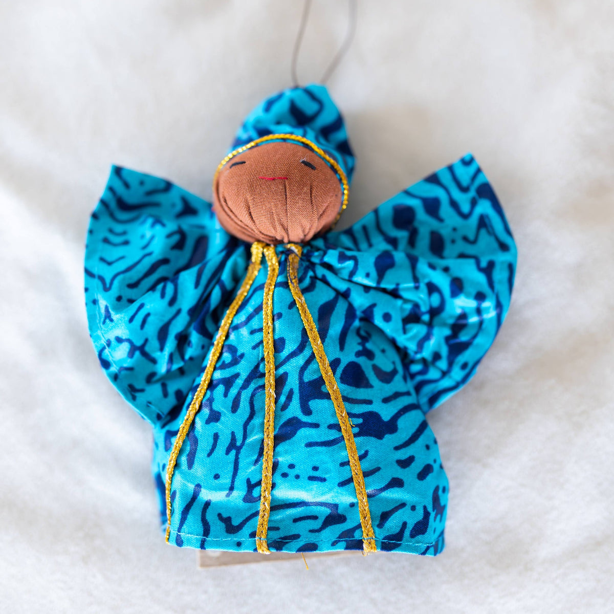 A handmade fabric angel ornament with bright blue and black patterned wings, a brown fabric head, and gold embroidered details, hanging against a white textured background.