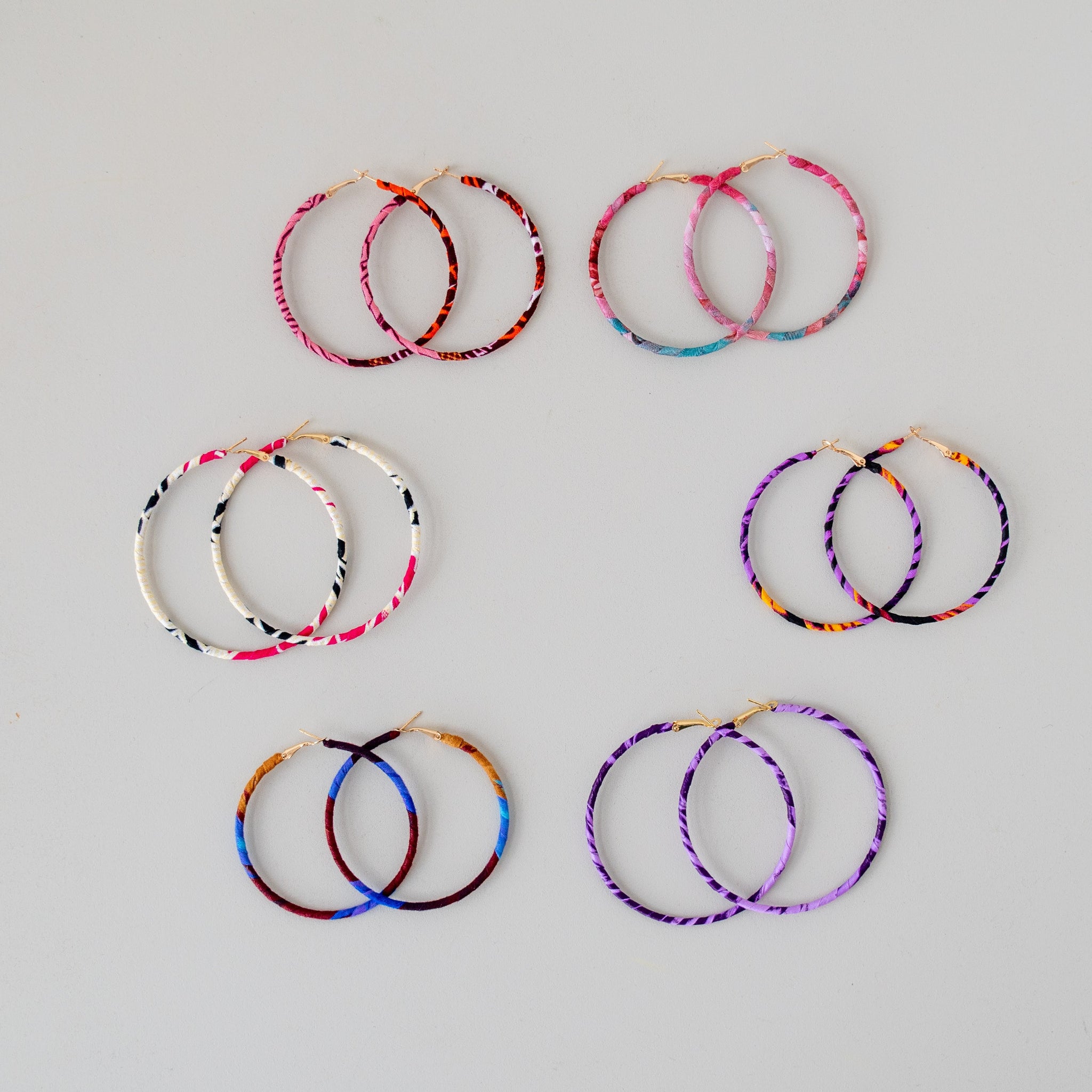 Seven pairs of large hoop earrings with colorful spiral patterns, arranged on a light gray background.