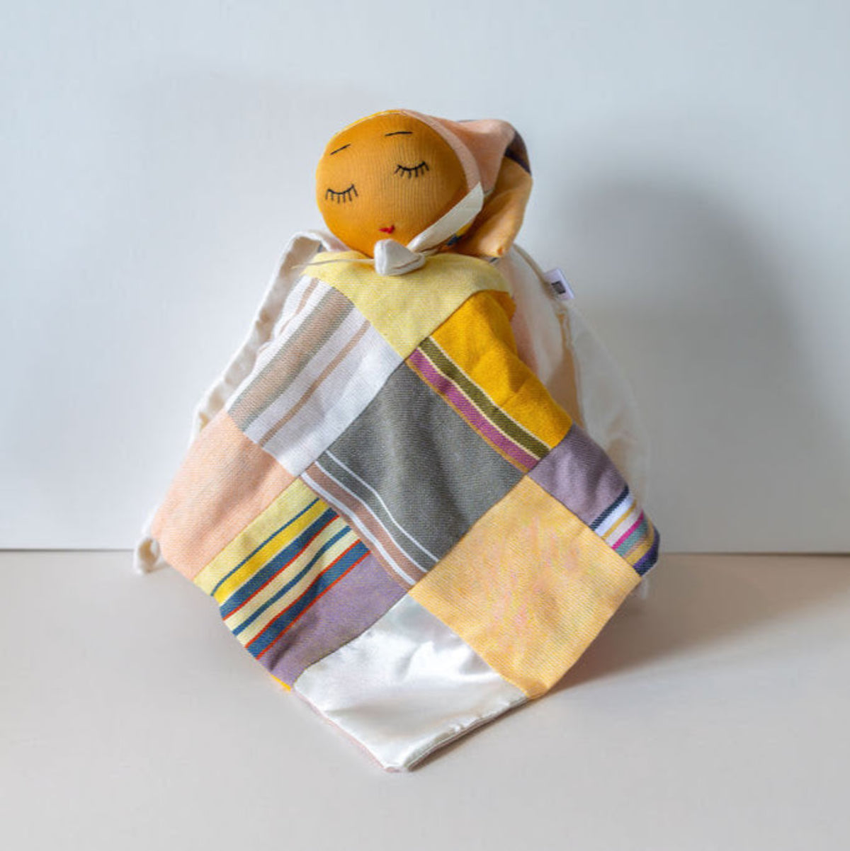 A handmade baby comforter featuring a smiling orange fabric face on a white cord, wrapped in a colorful patchwork quilt of yellow, gray, pink, and white fabrics, set against a plain light gray background.