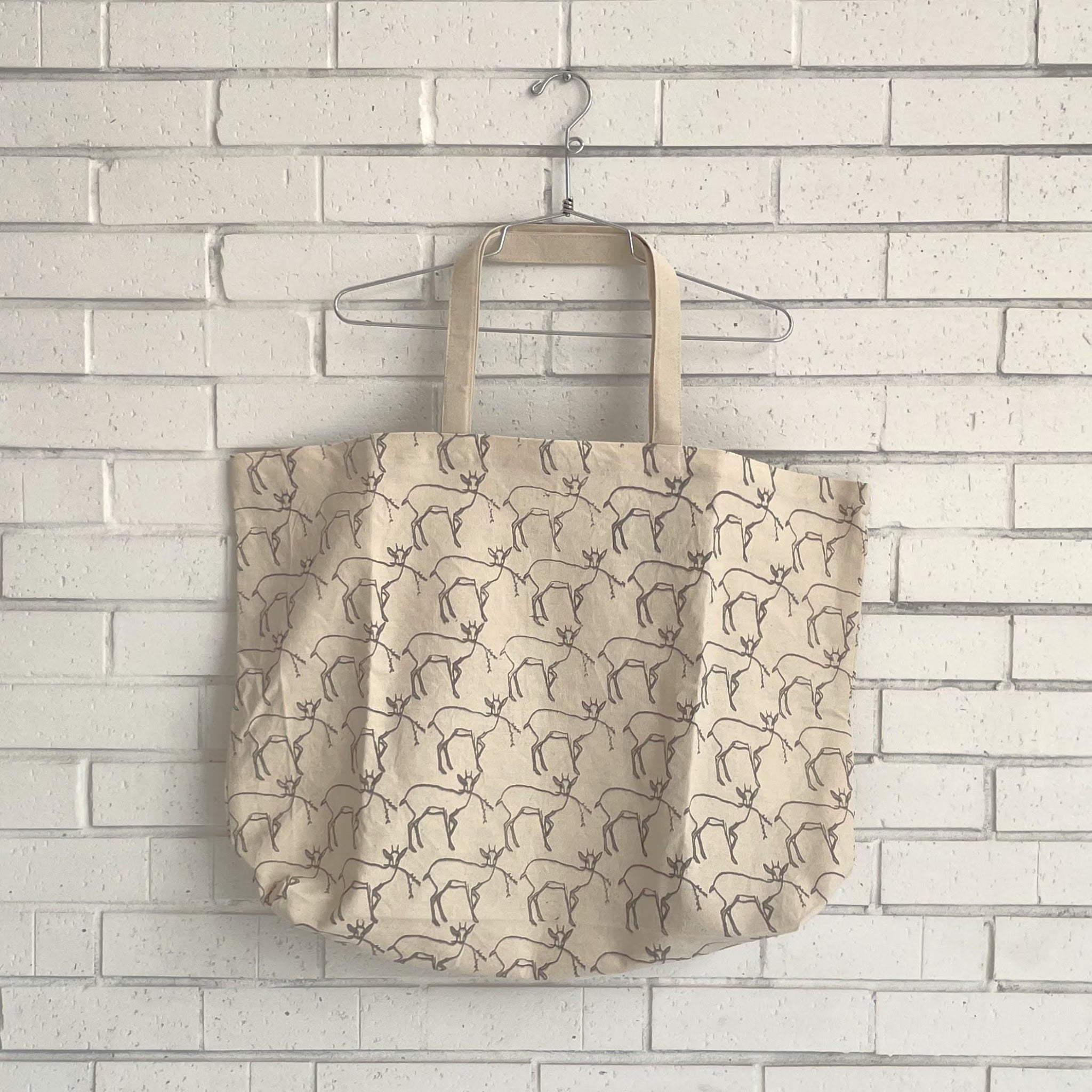 Beige tote bag with deer pattern hanging on a hanger against a light brick wall.