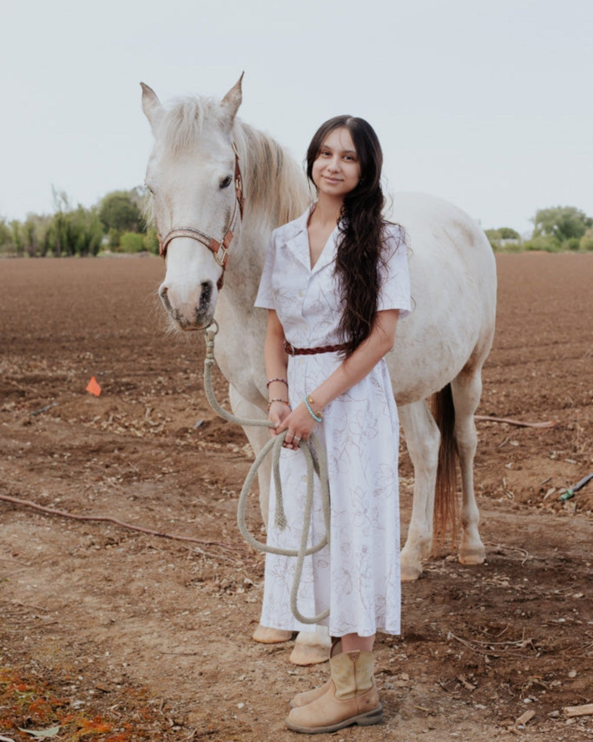 A person standing next to a white horse, wearing a white Magnolia Dress with a V-neck neckline.