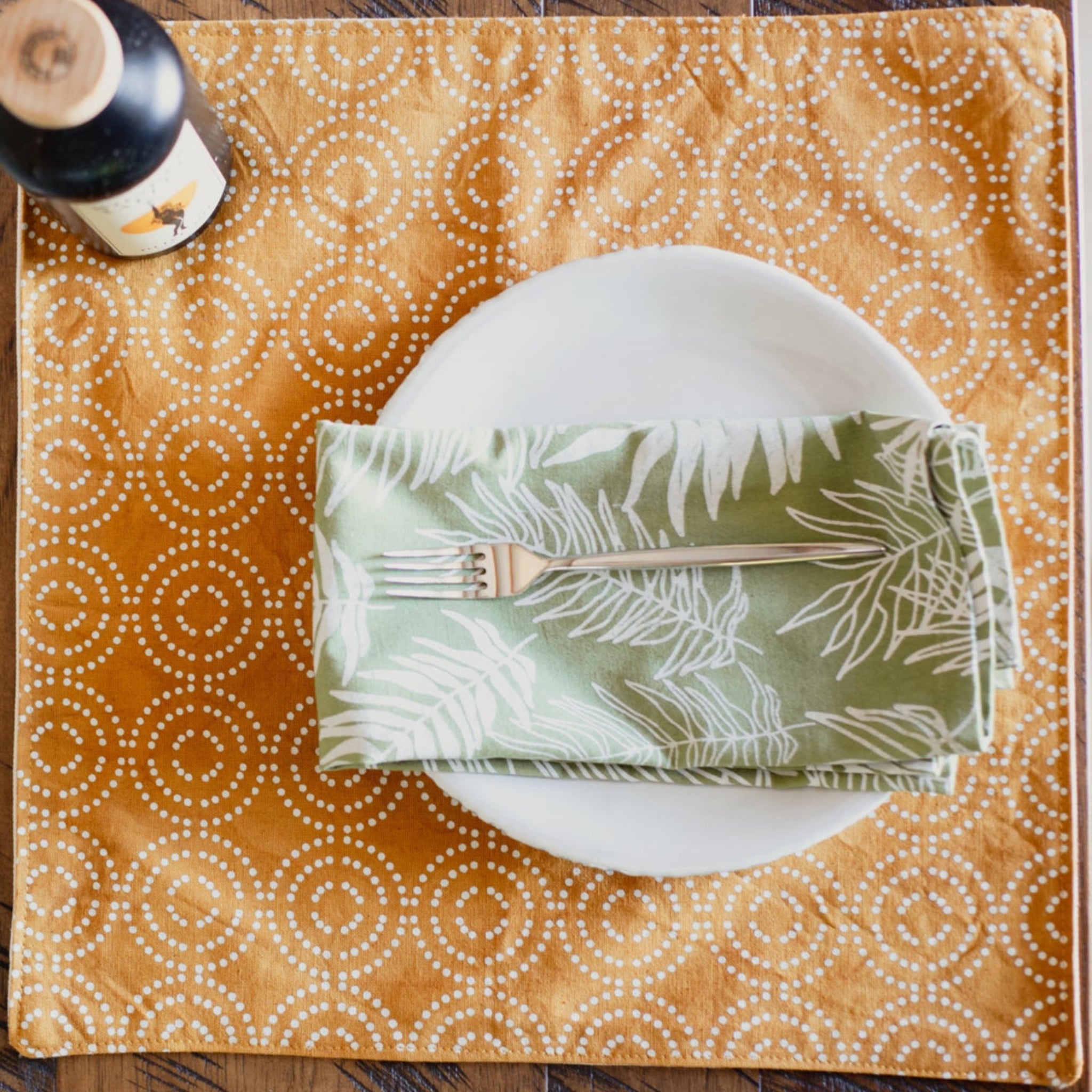 Placemat with circular pattern, white plate, green leaf-patterned napkin, and wine bottle.