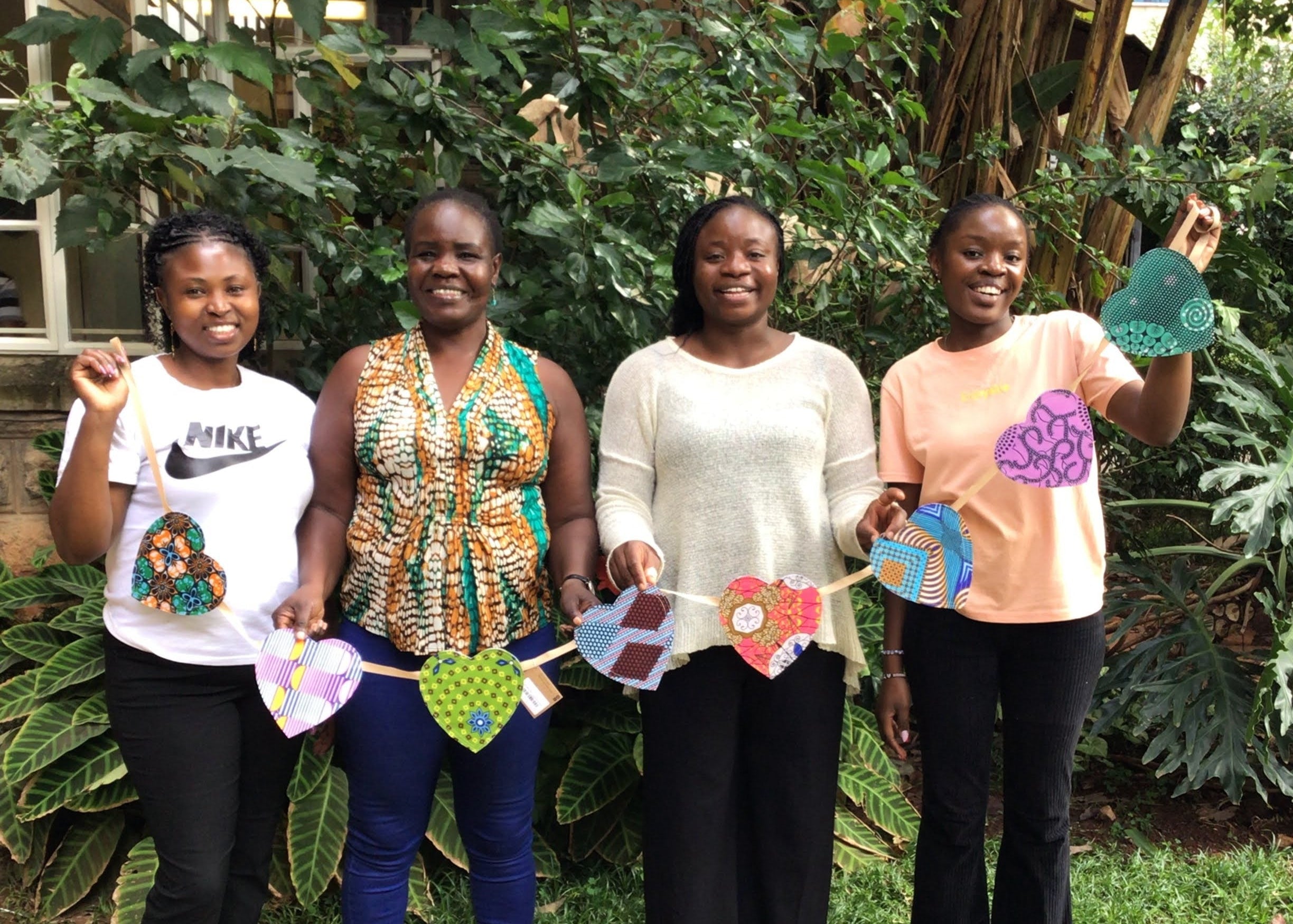 Four women standing outdoors with decorative items, surrounded by greenery.