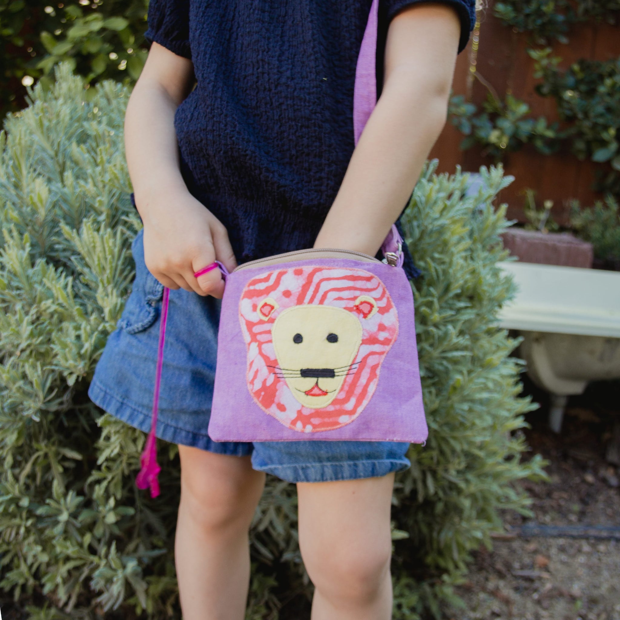 Child holding a purple pouch with a lion design in front of green bushes