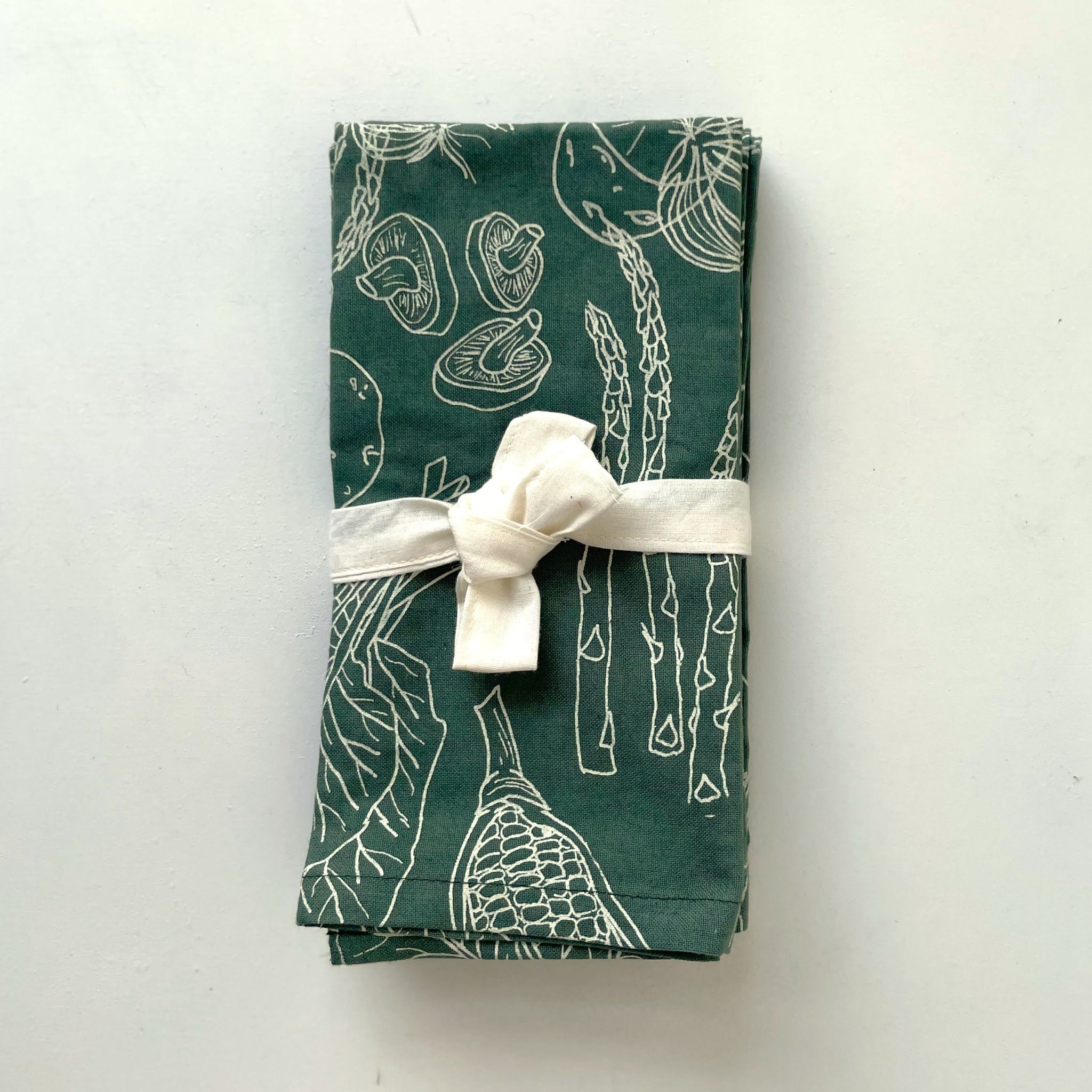 Green fabric napkins with white line drawings of vegetables tied with a white ribbon on a white background