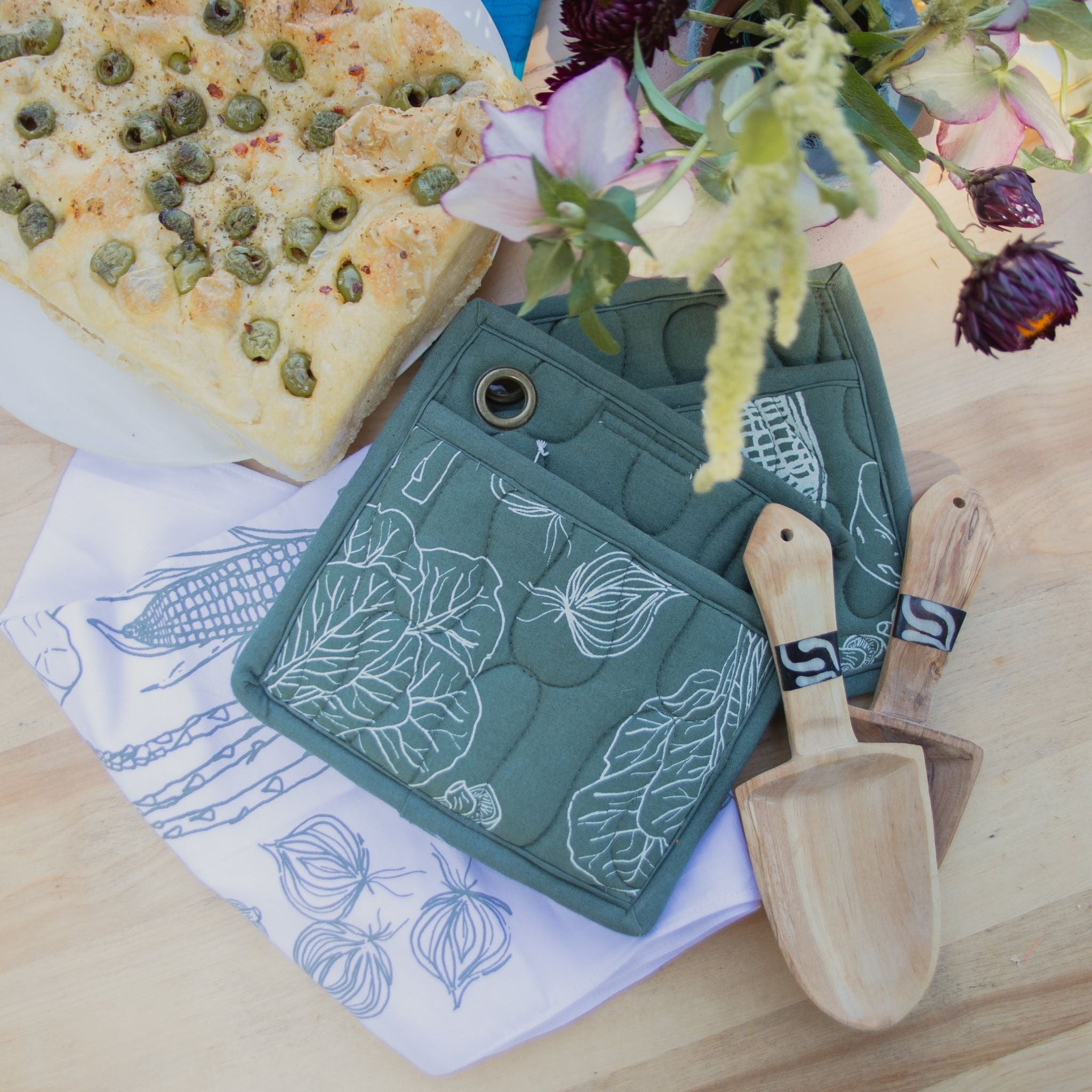 Green pot holder with floral design on a wooden table with focaccia bread and flowers.
