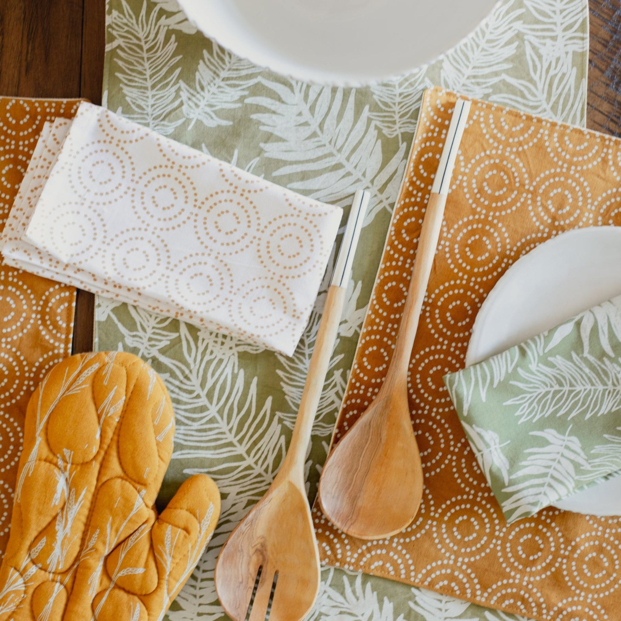 Table setting with patterned tablecloth, wooden spoons, and oven mitts on a wooden surface.