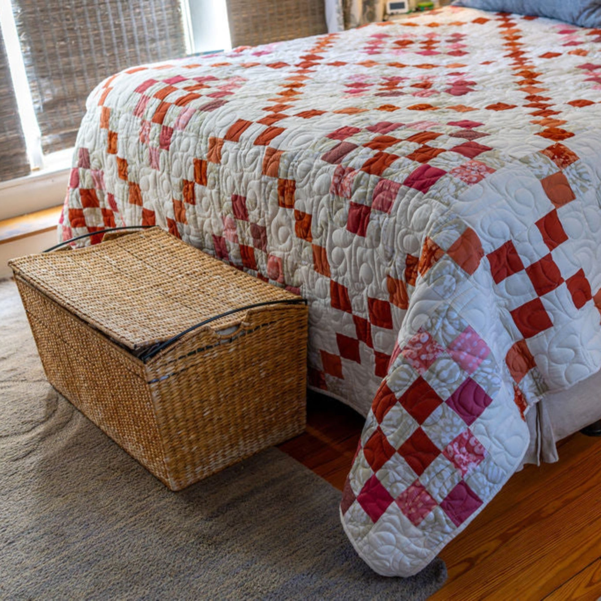 A quilted patchwork bedspread with red, pink, and orange diamond blocks on a white background, styled on a bed in a bedroom with a woven storage basket at the foot.