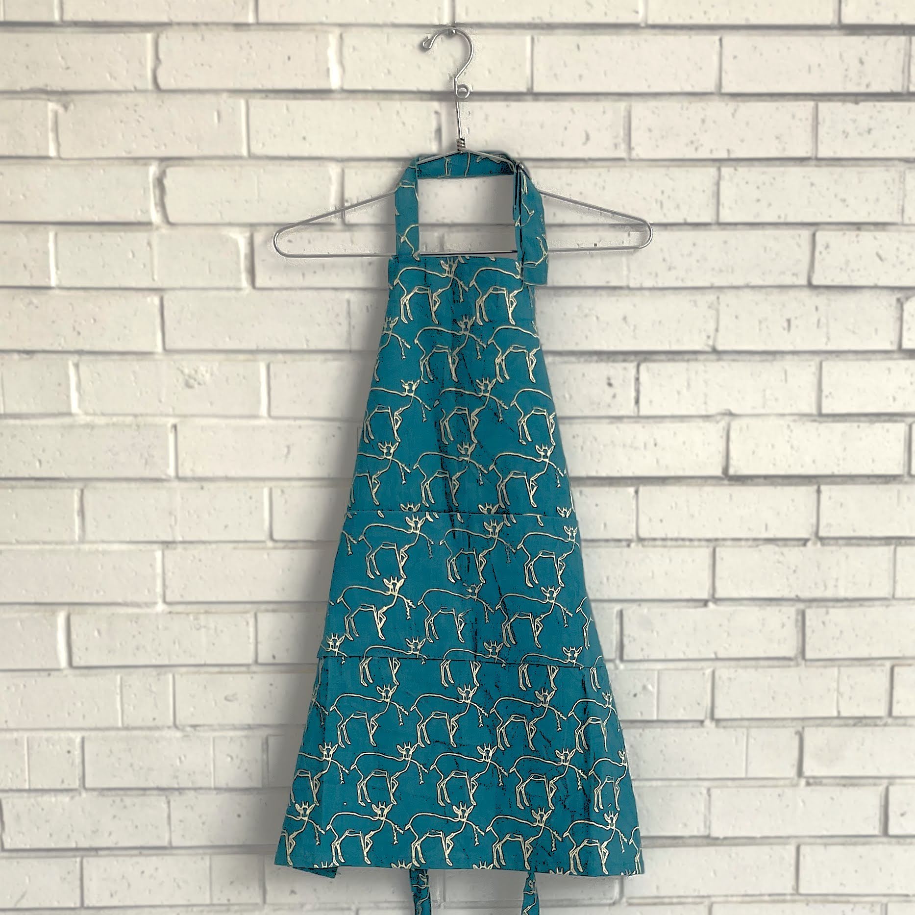 Teal apron with gold pattern on a hanger against a white brick wall.