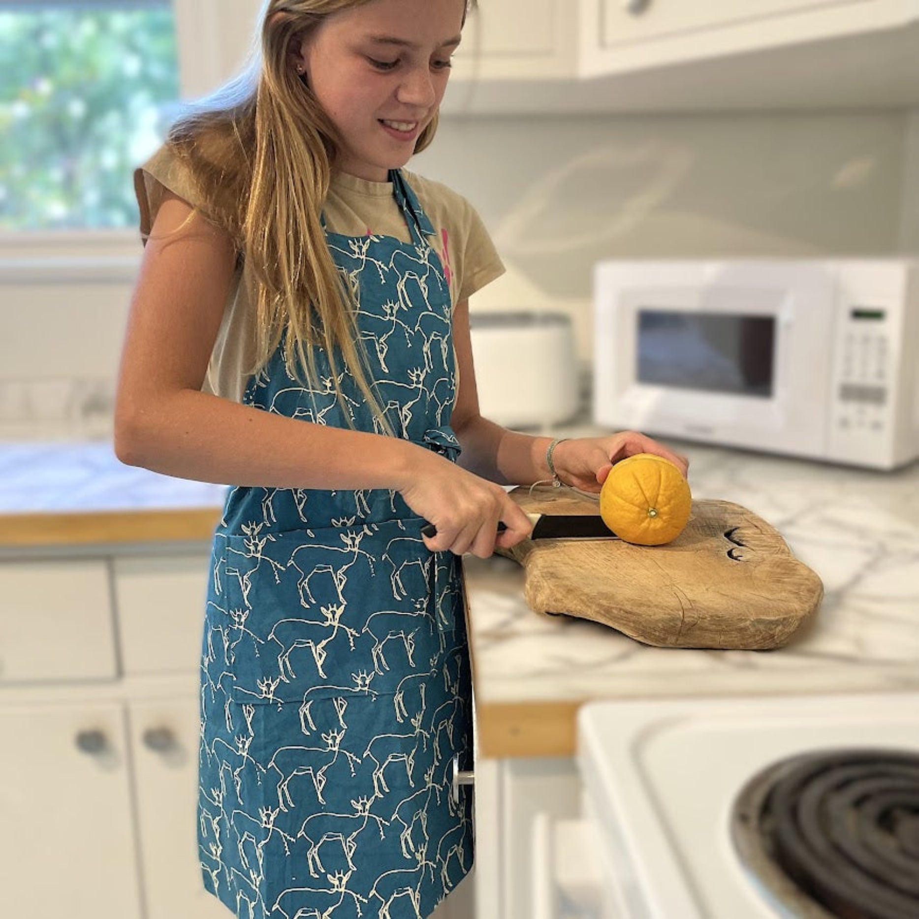 Young girl in a kitchen wearing a blue apron with a white pattern, cutting an orange on a wooden cutting board.