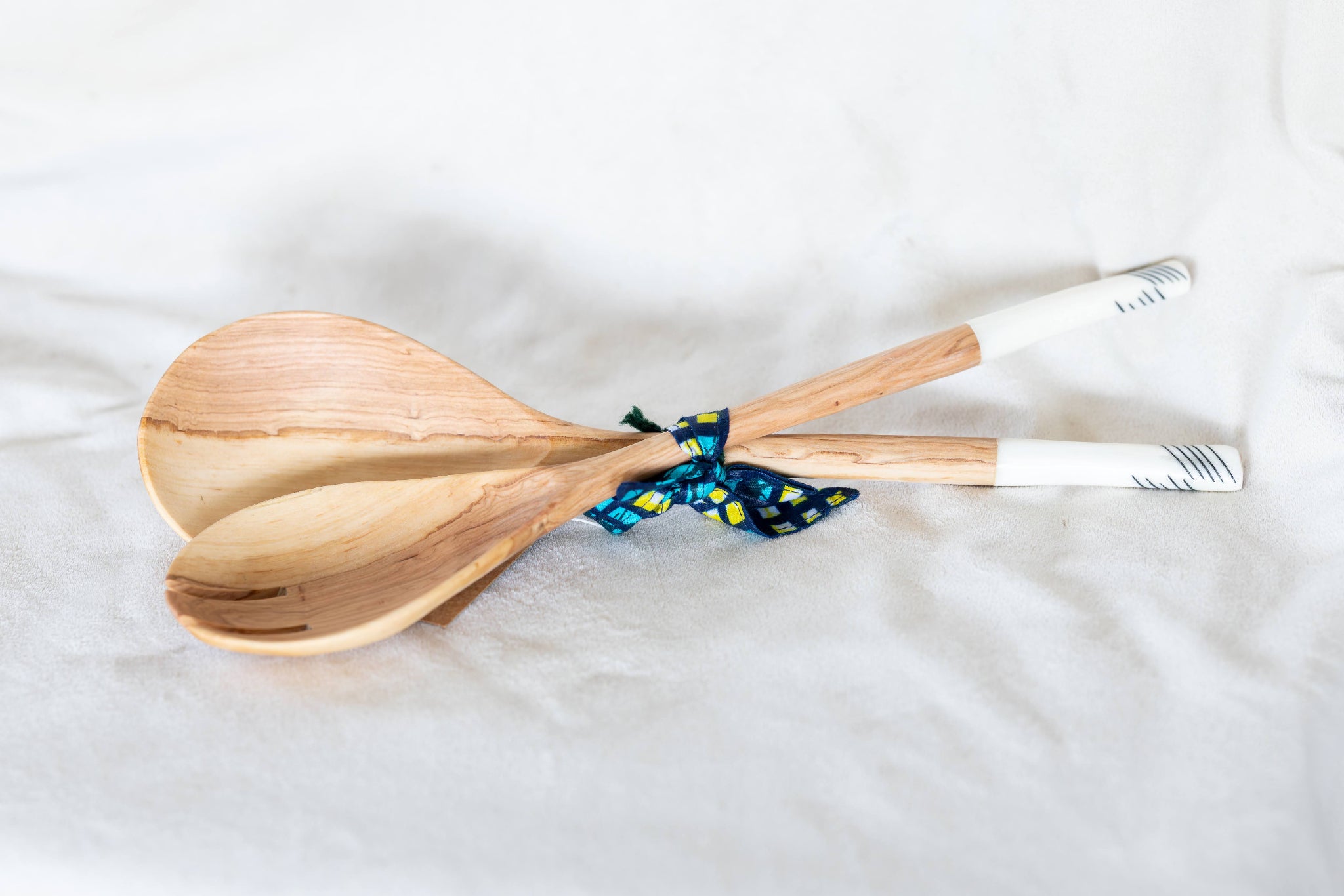 Two light natural wood serving spoons tied together with a blue and yellow patterned ribbon, resting on a white fabric background.