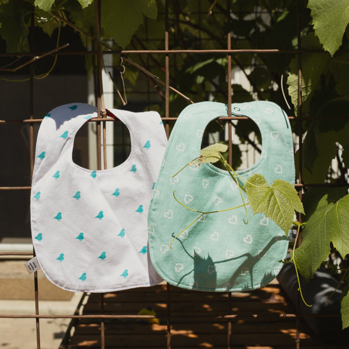 Two baby bibs with leaf patterns hanging on a metal rack outdoors.