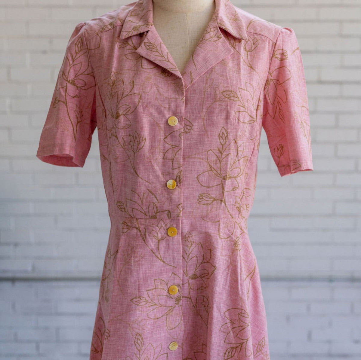Detail of a pink button up dress with tan floral pattern in front of white brick wall.