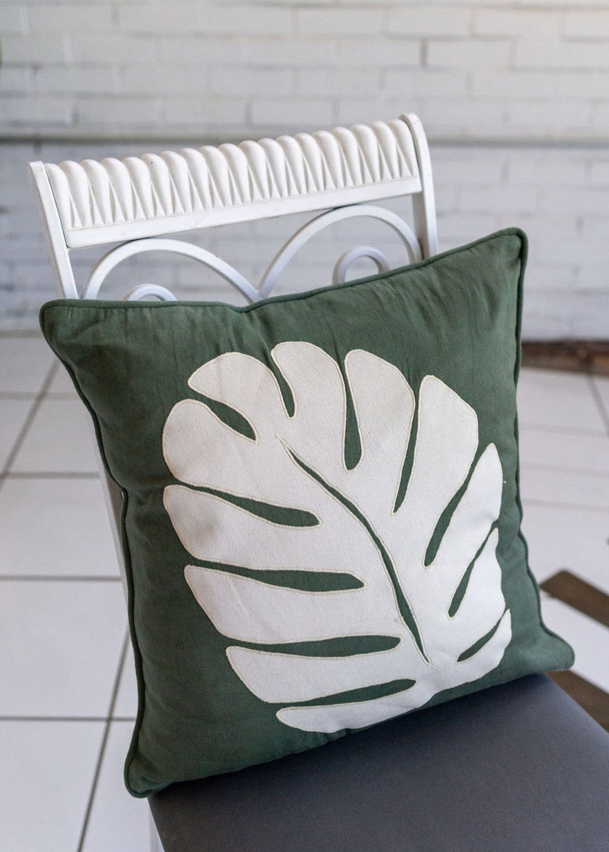 A green pillow with a cream monstera leaf on a white and gray chair in front of a white brick wall