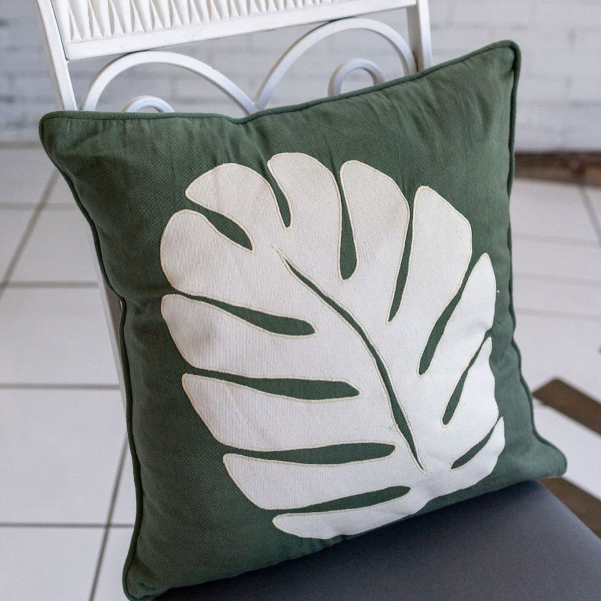 A green pillow with a cream monstera leaf on a white and gray chair in front of a white brick wall
