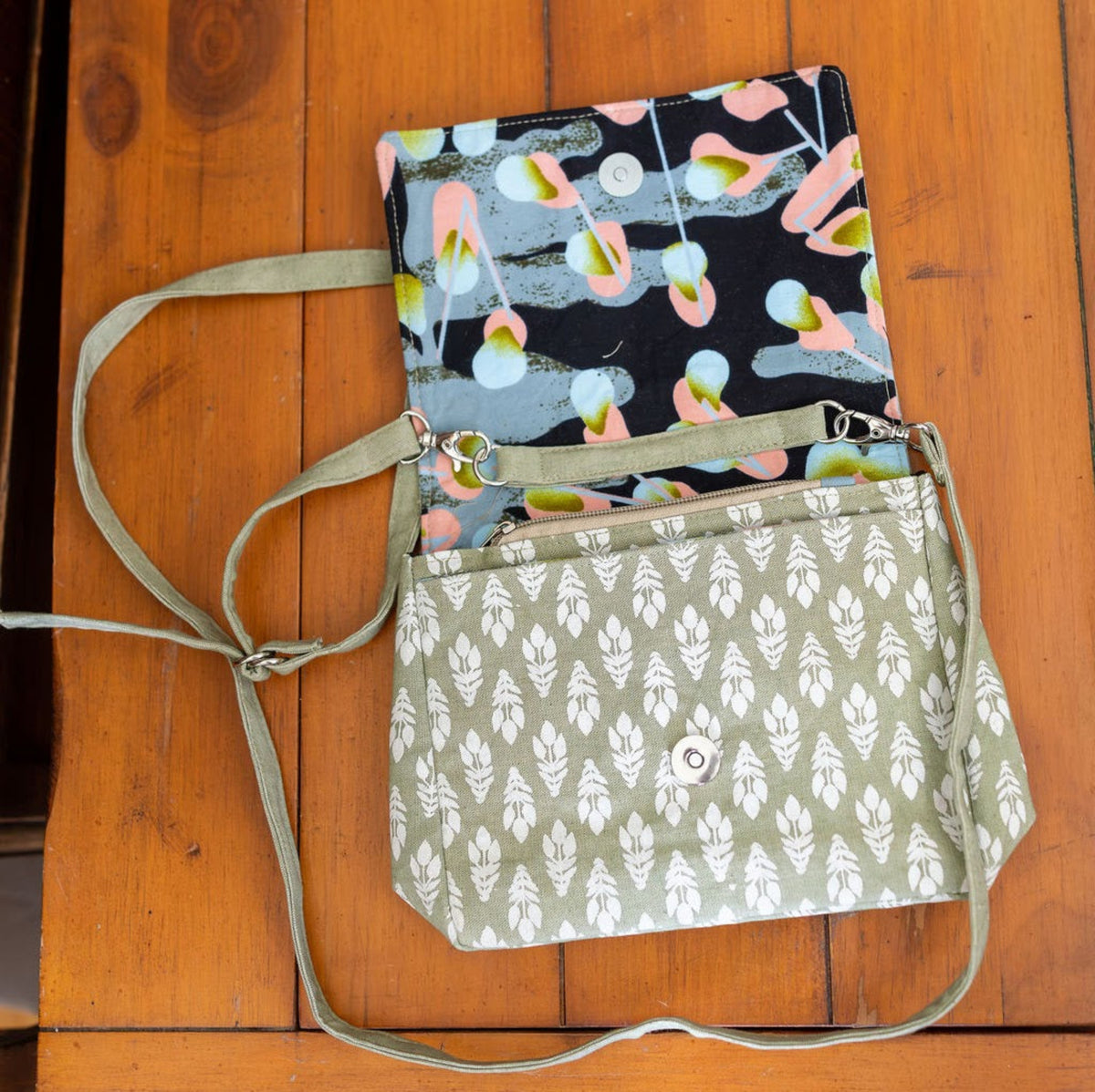 Opened green floral crossbody bag showing black floral inside. Bag is on a wooden table.