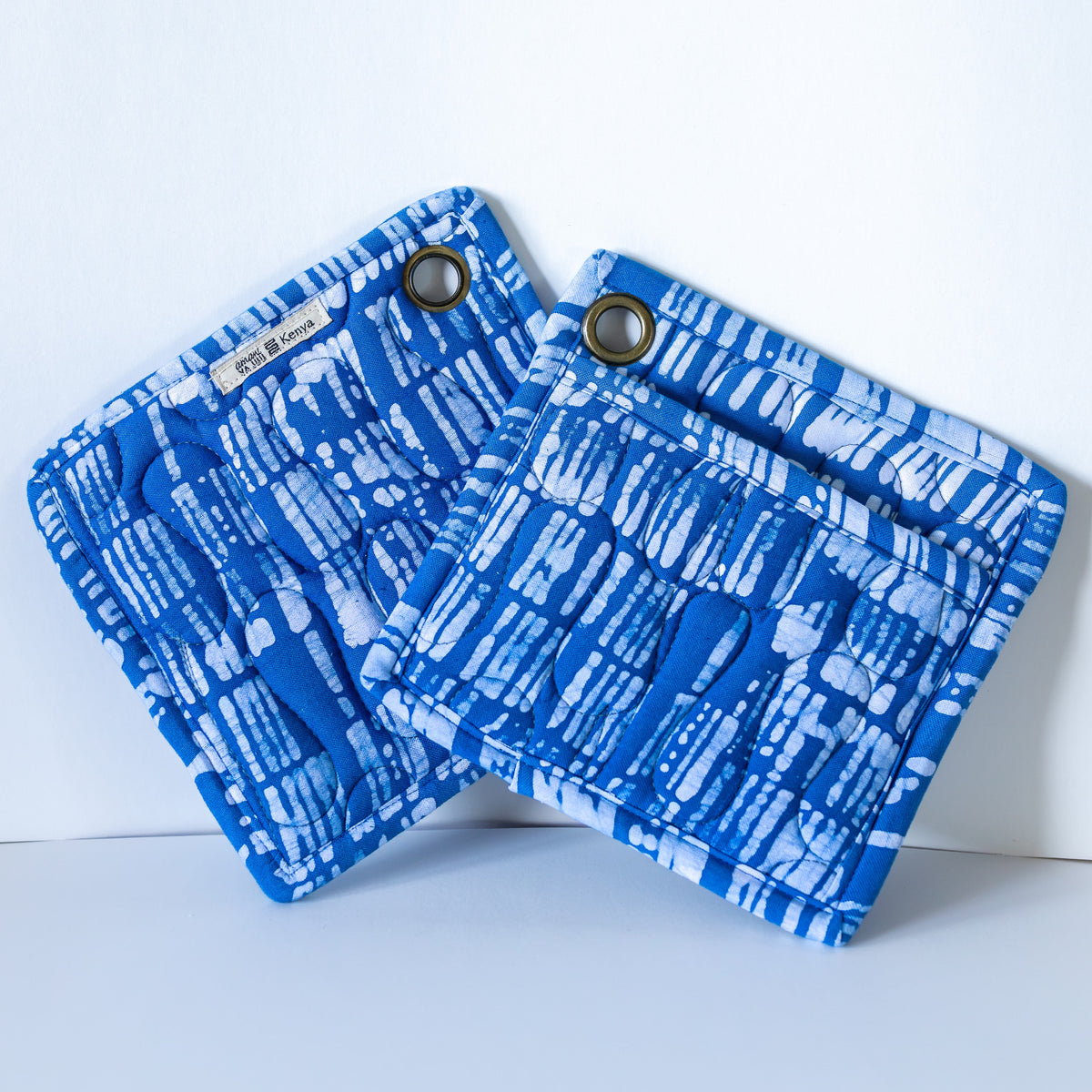 Two blue and white patterned quilted potholders with metal grommets, displayed on a clean white background.