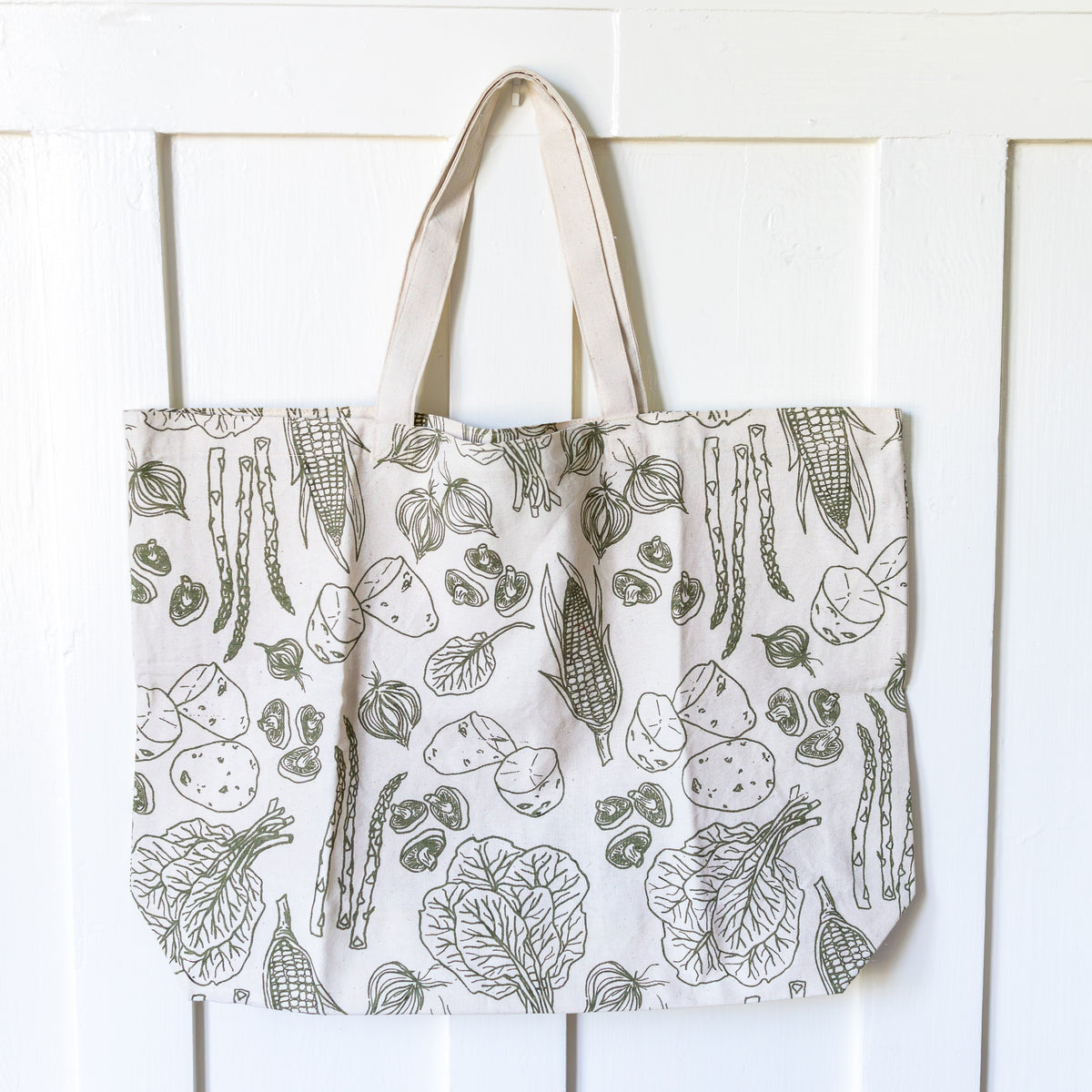 Large cream tote bag with olive green vegetable pattern hanging on a white door