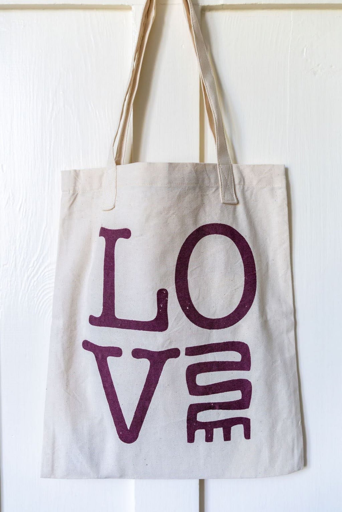 A natural canvas tote bag hanging against a white paneled wall, featuring the word 'LOVE' printed in a stylized dark purple font.
