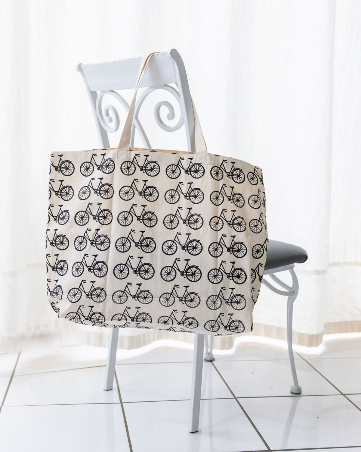 Large natural colored tote bag with black bicycle print hanging from a white chair in a white room.