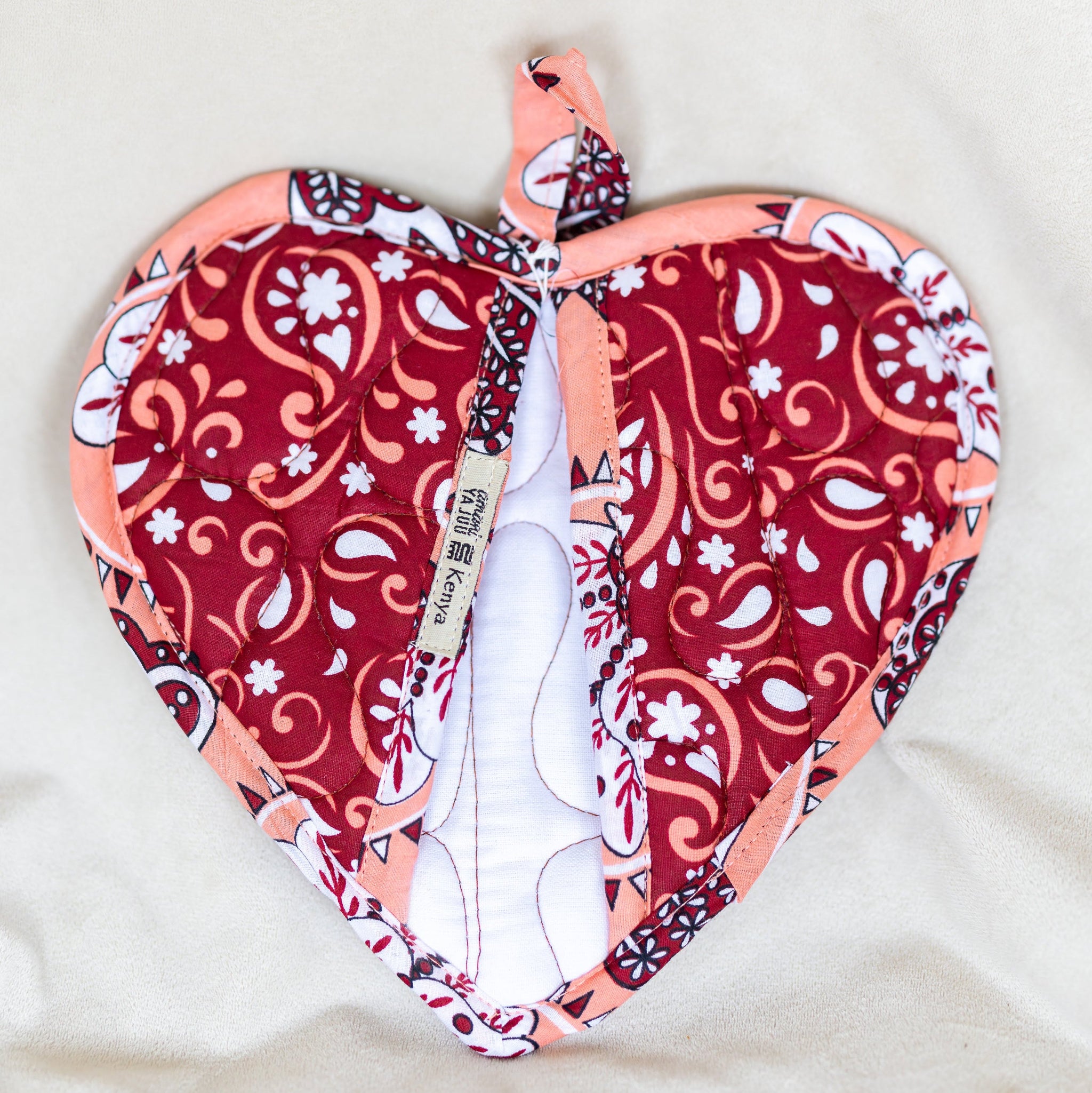 A heart-shaped quilted fabric potholder with a red, white, and peach paisley pattern, laid flat on a light beige fabric background.