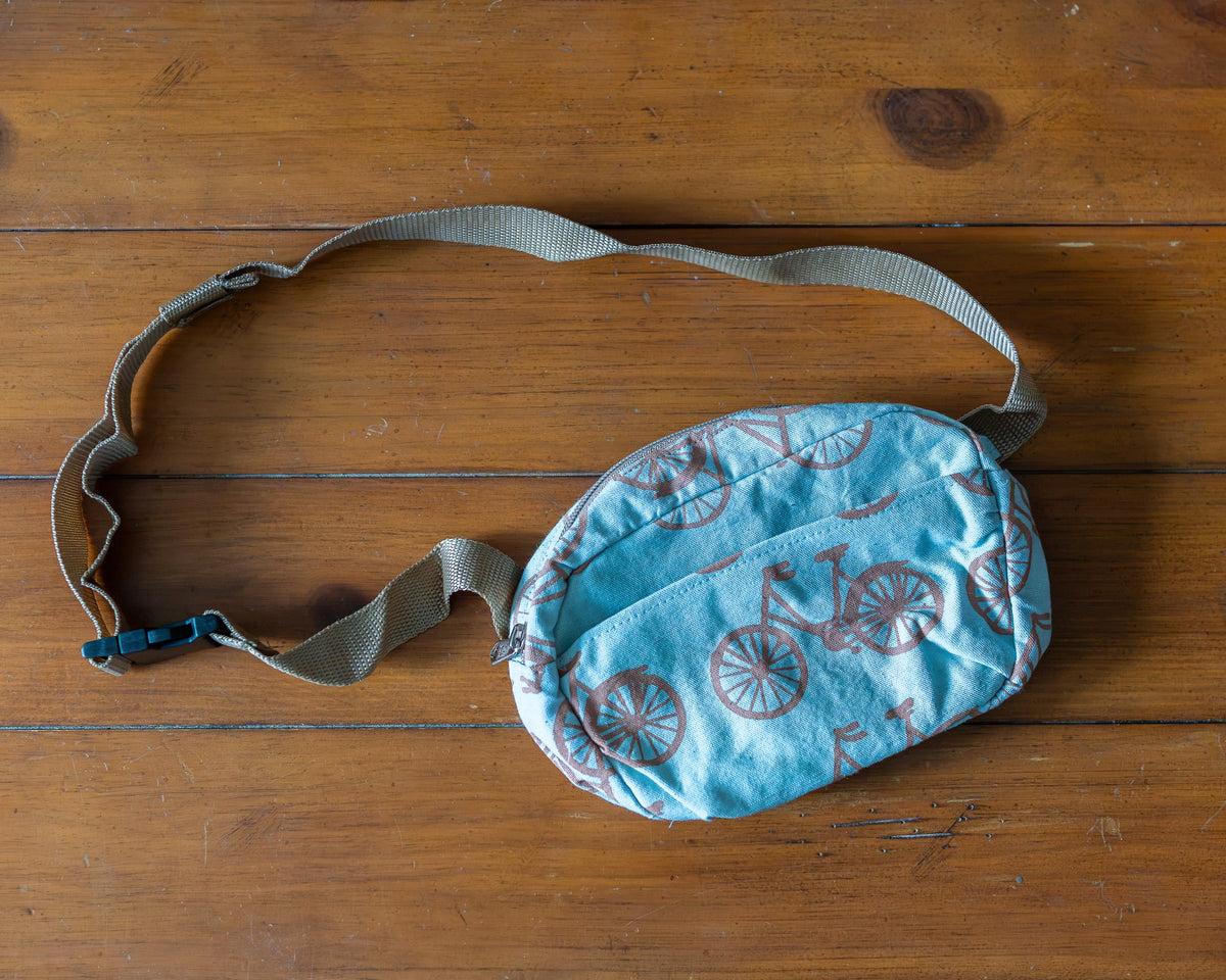 A small light blue fabric pouch with a brown bicycle pattern, featuring an adjustable beige webbing strap and a quick-release plastic buckle, laid flat on a wooden surface.