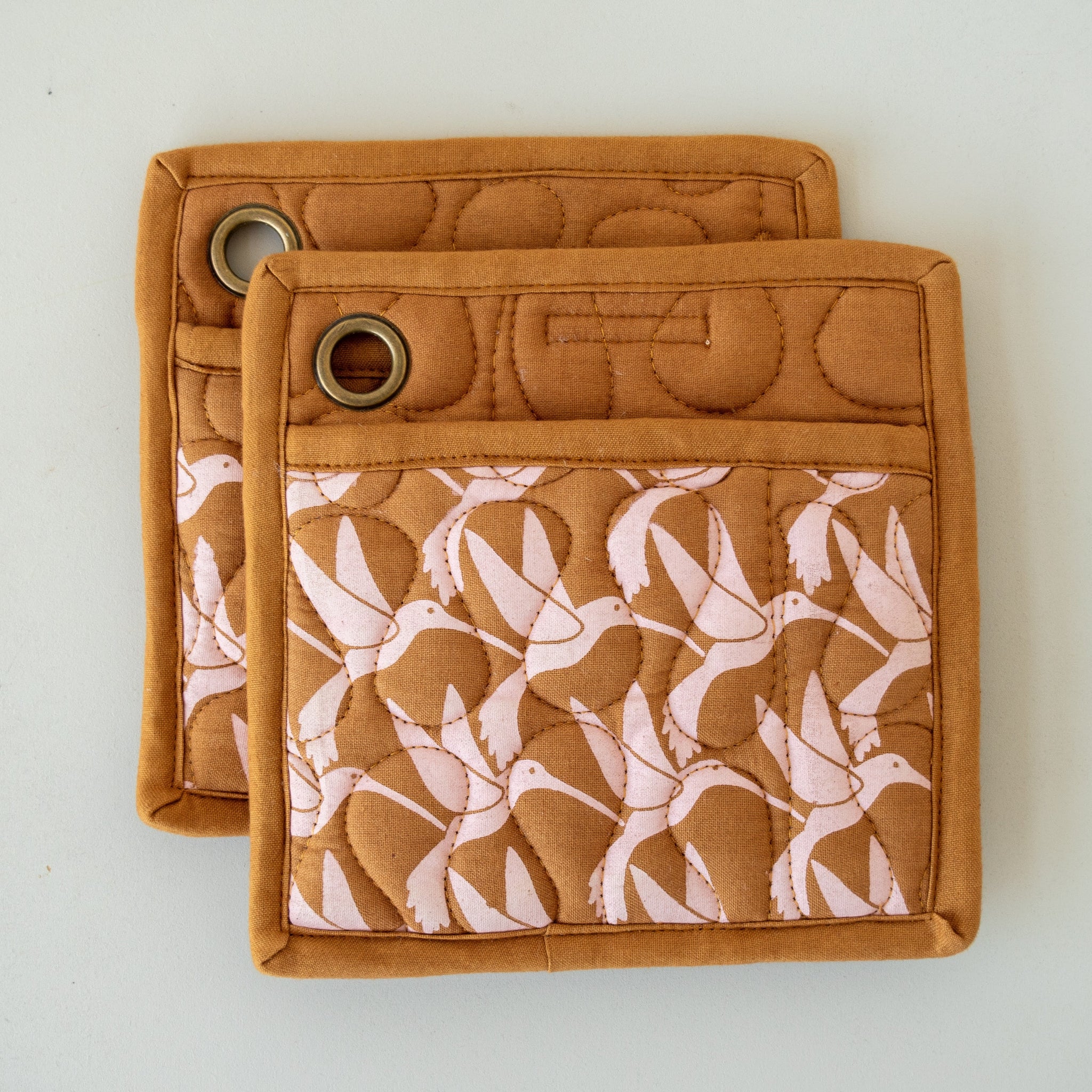 Two brown square pot holders with white bird screen print and a pocket on each, with metal loops for hanging