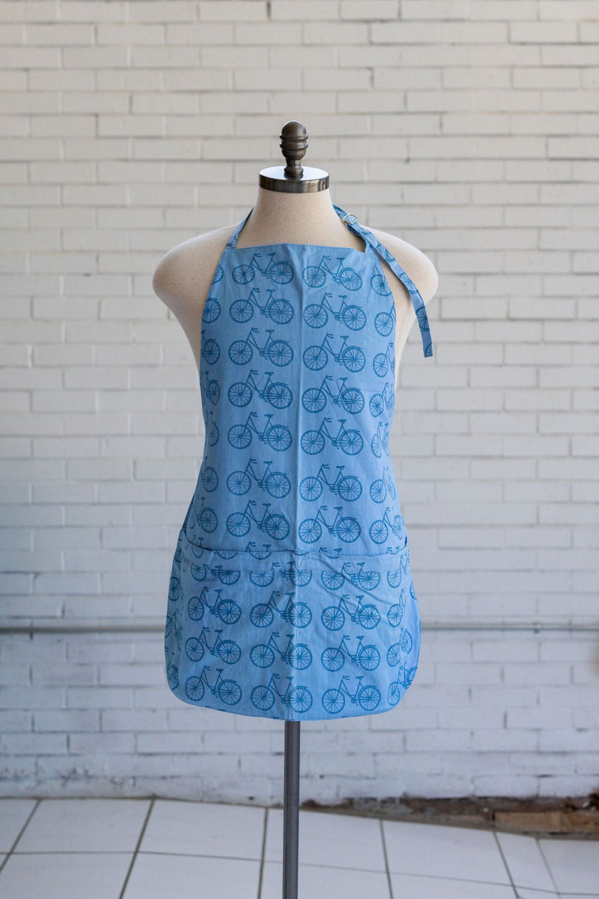 A light blue halter-style apron with a repeating bicycle print, displayed on a mannequin torso in front of a white brick wall.