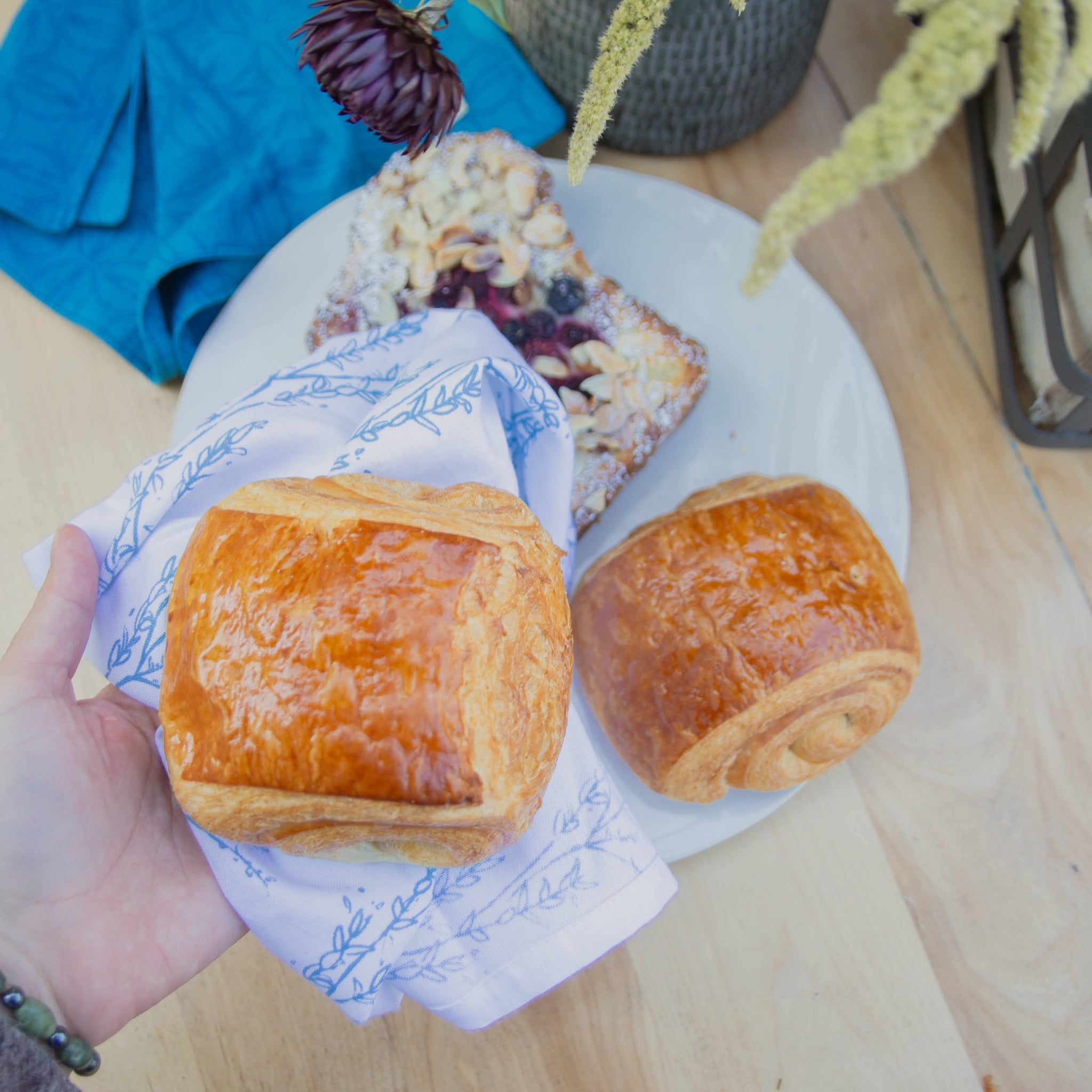 Two pastries on a white plate and one held by a napkin in hand with a plant in the background.