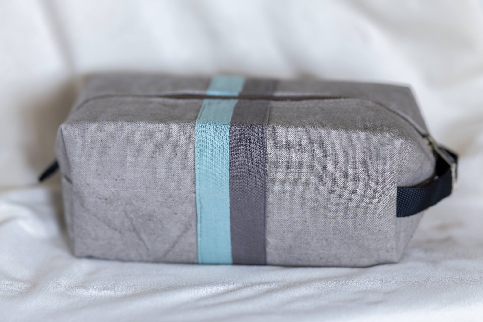 A gray fabric toiletry bag with a vertical striped pattern in light blue, dark gray, and black, resting on a white fabric surface.