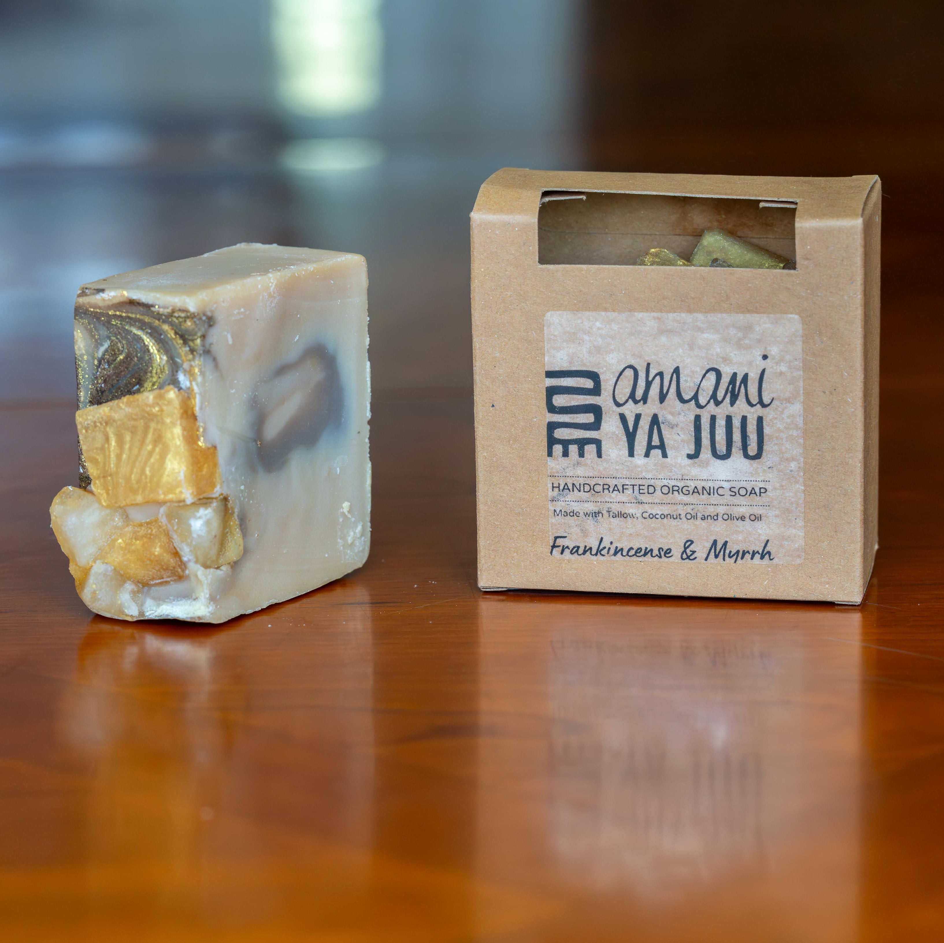 A handmade organic soap bar with gold and blue marbled swirls sits next to its kraft paper packaging box on a polished wooden table. The box is labeled 'Amani Ya Juu' and 'Frankincense & Myrrh'