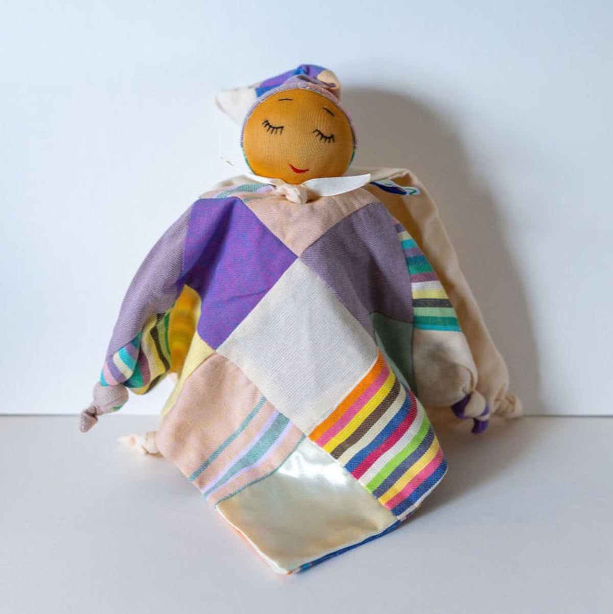 A handmade patchwork fabric doll with a round orange face, embroidered closed eyes, and a multicolor quilted body in purple, beige, green, and striped patterns, set against a plain light gray background.