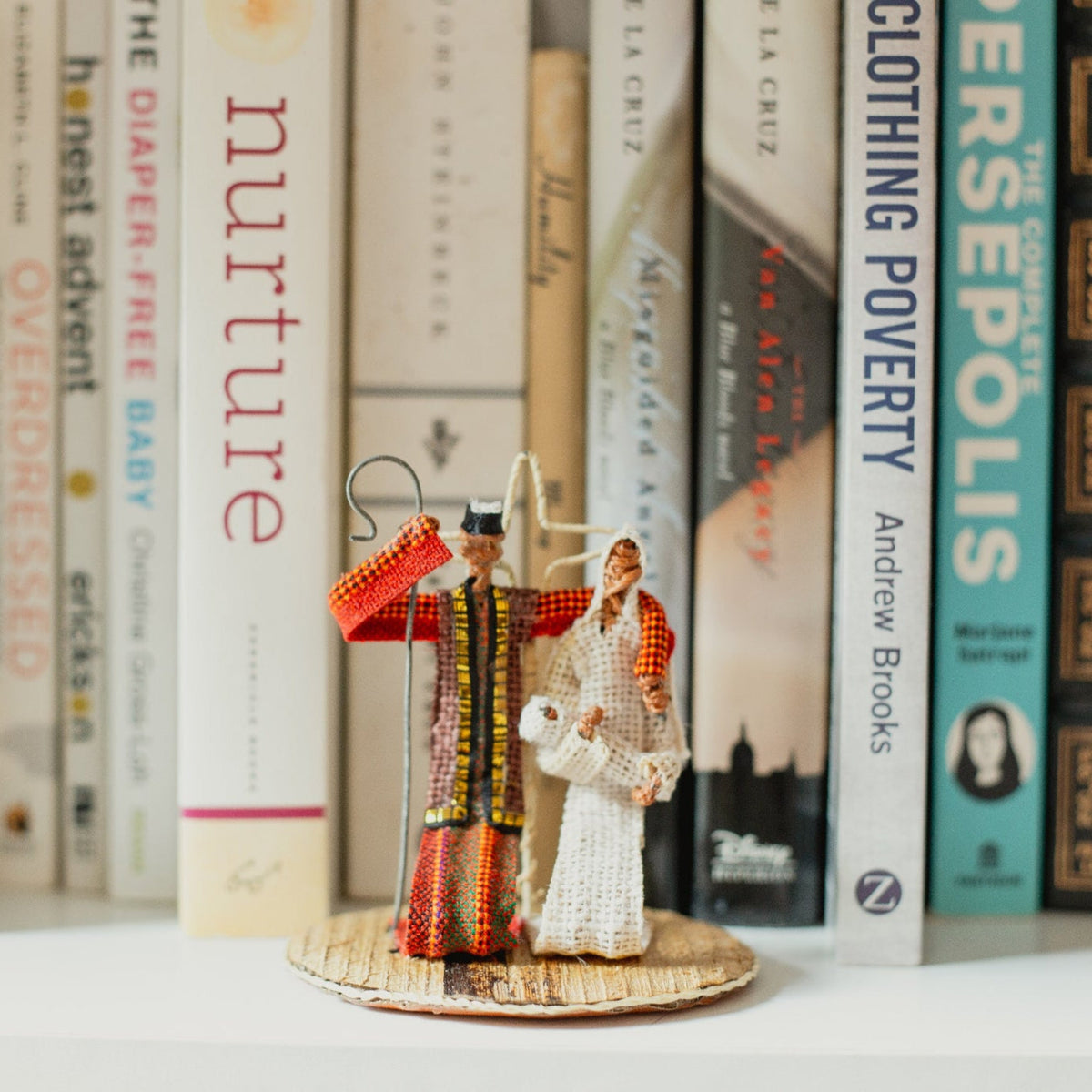 Three small figurines of the holy family on a bookshelf with books in the background.