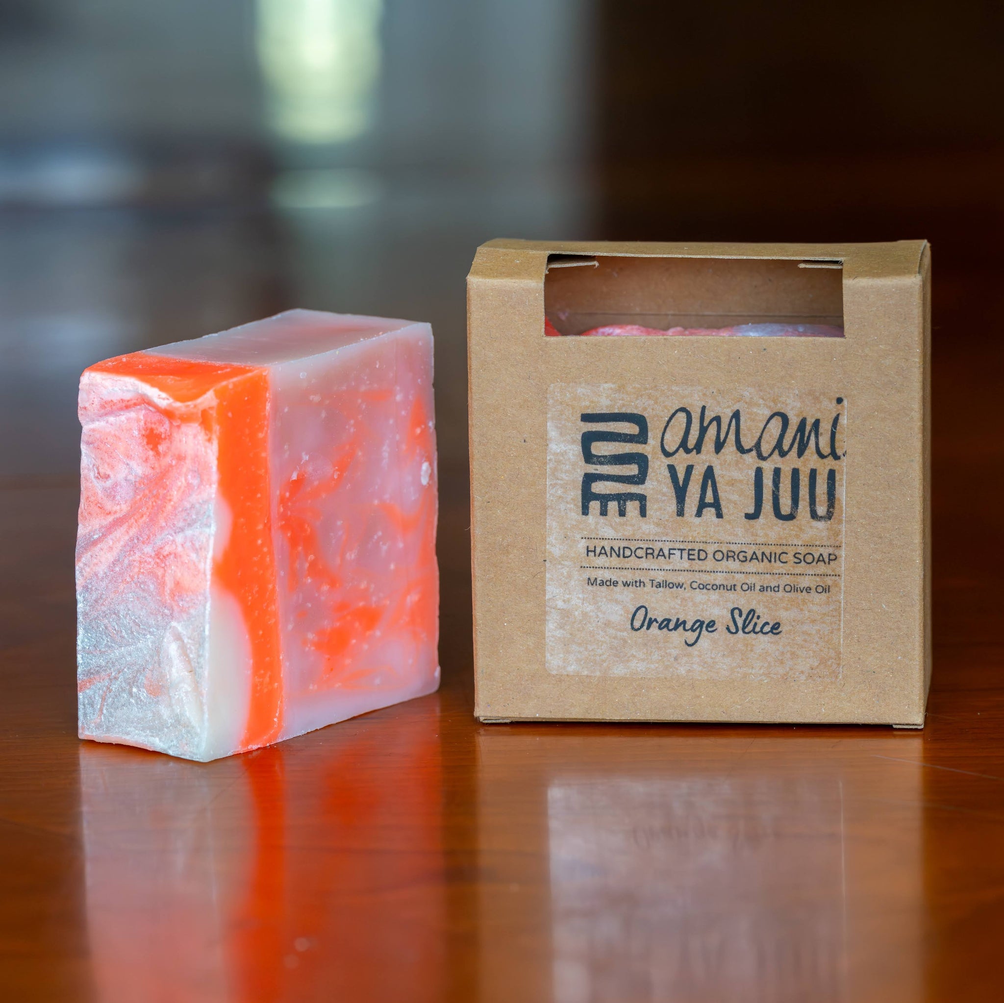 A rectangular bar of orange and white marbled soap stands beside a kraft paper box labeled 'amani YA JUU' and 'Orange Slice' on a polished wooden surface.