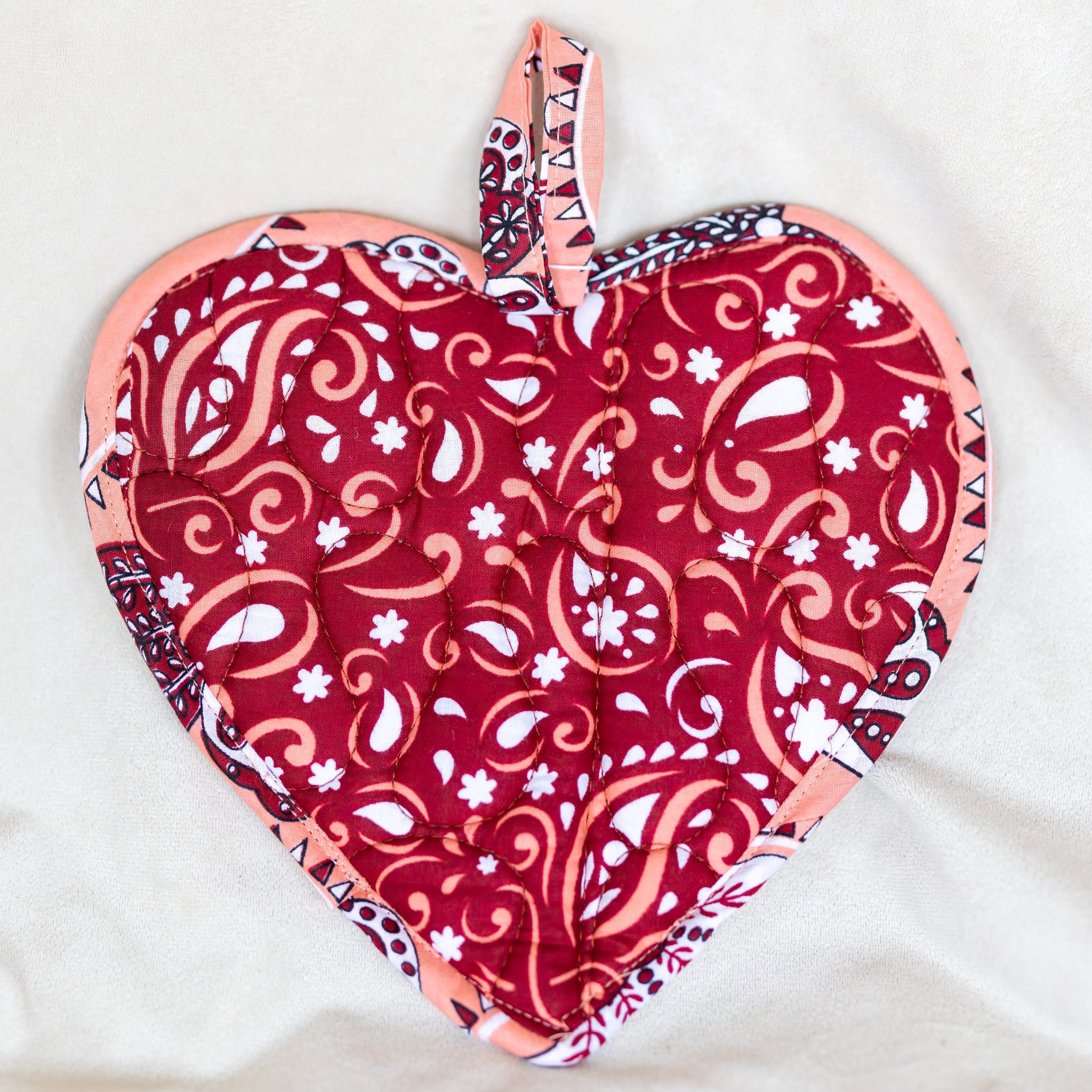 A heart-shaped quilted potholder with a red, pink, and white paisley pattern, featuring a matching fabric loop at the top, laid flat on a white fabric background.