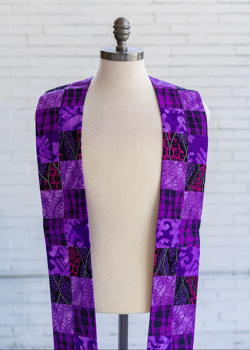 A purple patchwork stole displayed on a cream fabric mannequin torso, set against a white brick wall background.