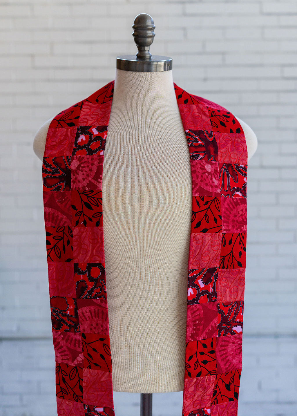 A red and black batik patchwork stole draped on a cream fabric mannequin torso, set against a white brick wall background.