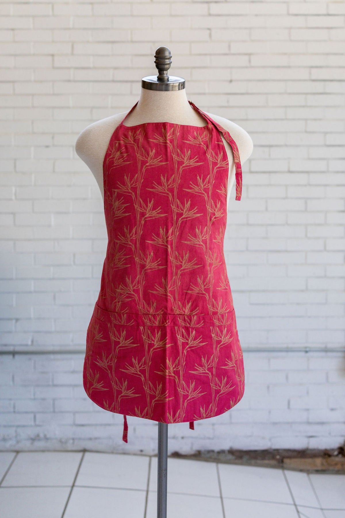 A pink halter-style apron with a gold bamboo pattern, displayed on a mannequin torso against a white brick wall background