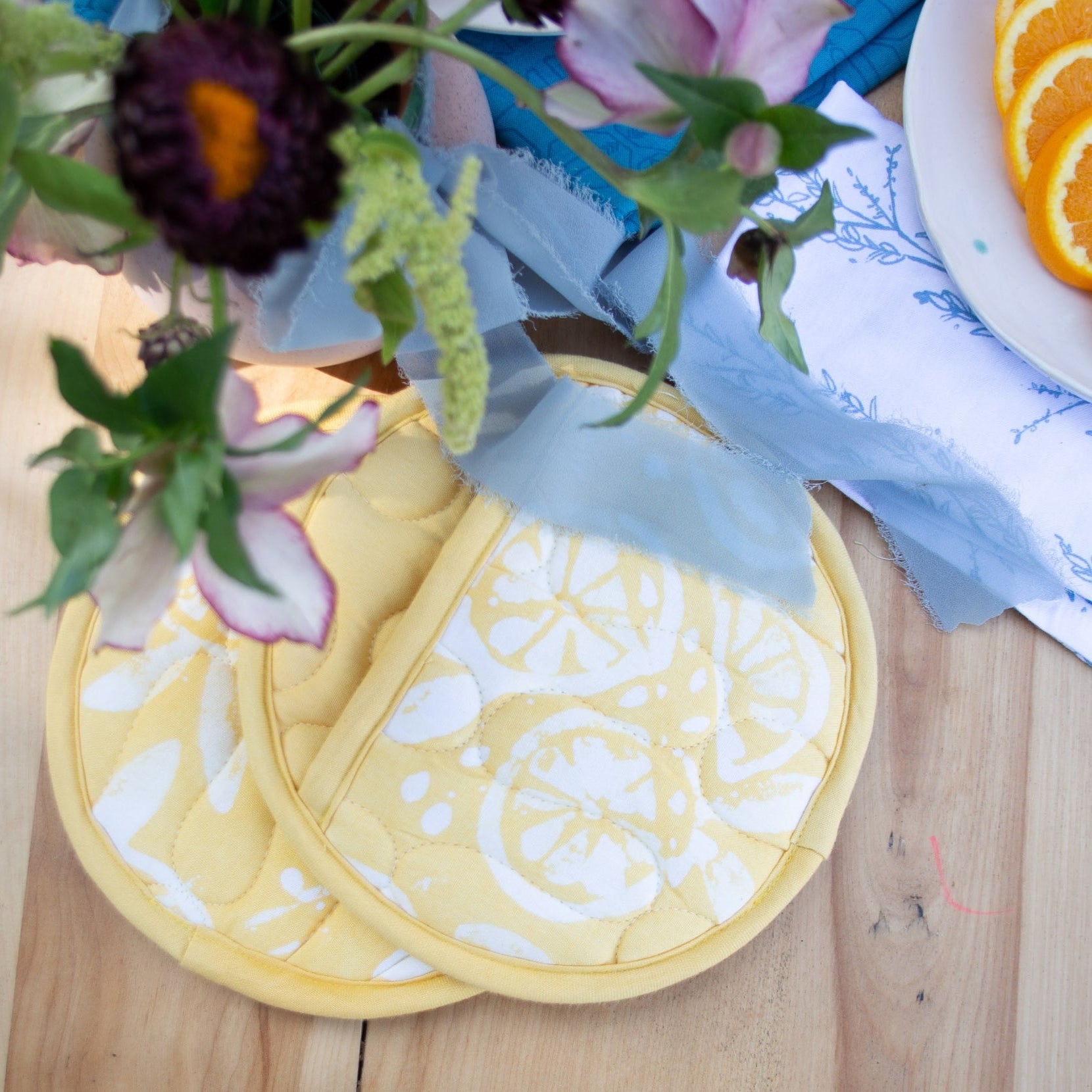 Yellow floral coasters on a wooden surface with a plate of oranges and flowers.