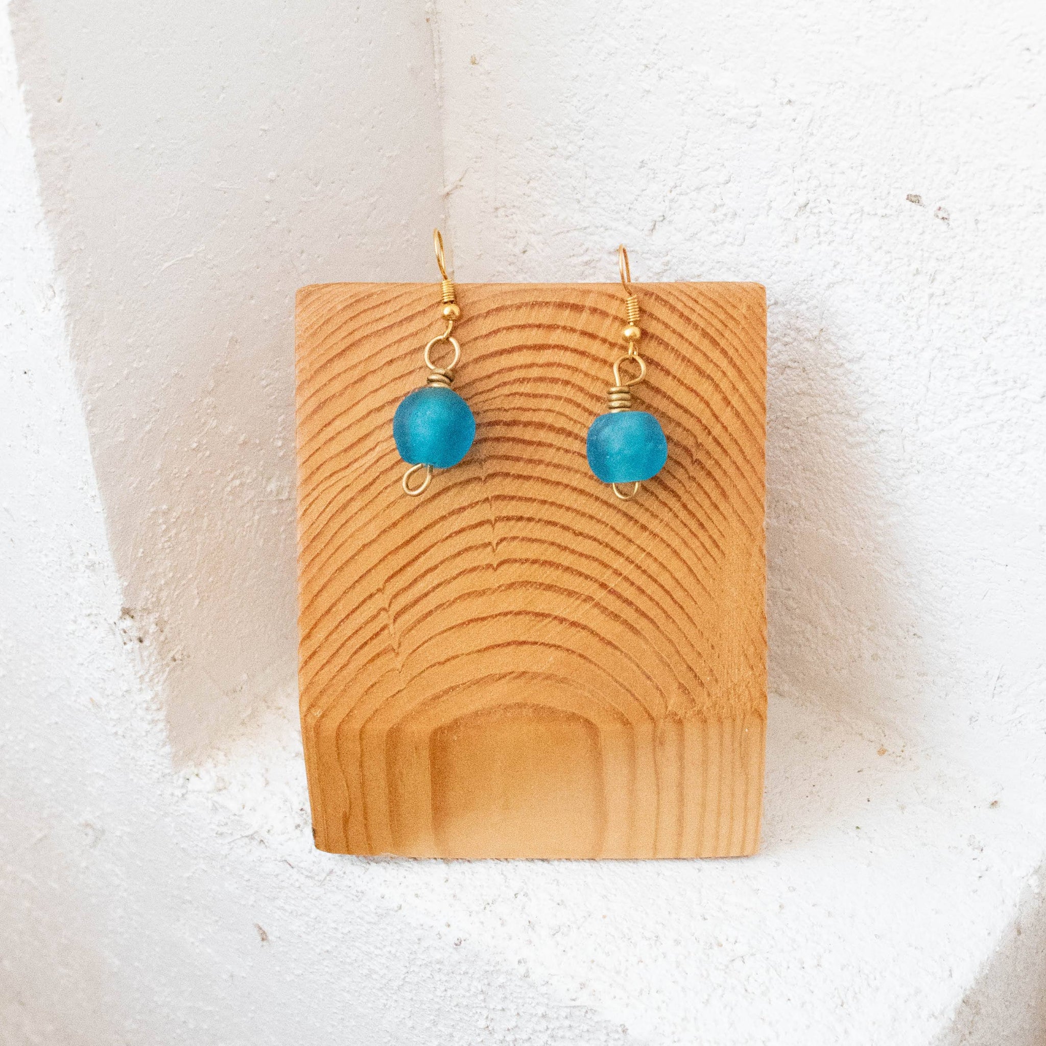 A pair of gold-tone dangle earrings with round turquoise-blue beads, displayed on a small wooden stand against a white textured wall.