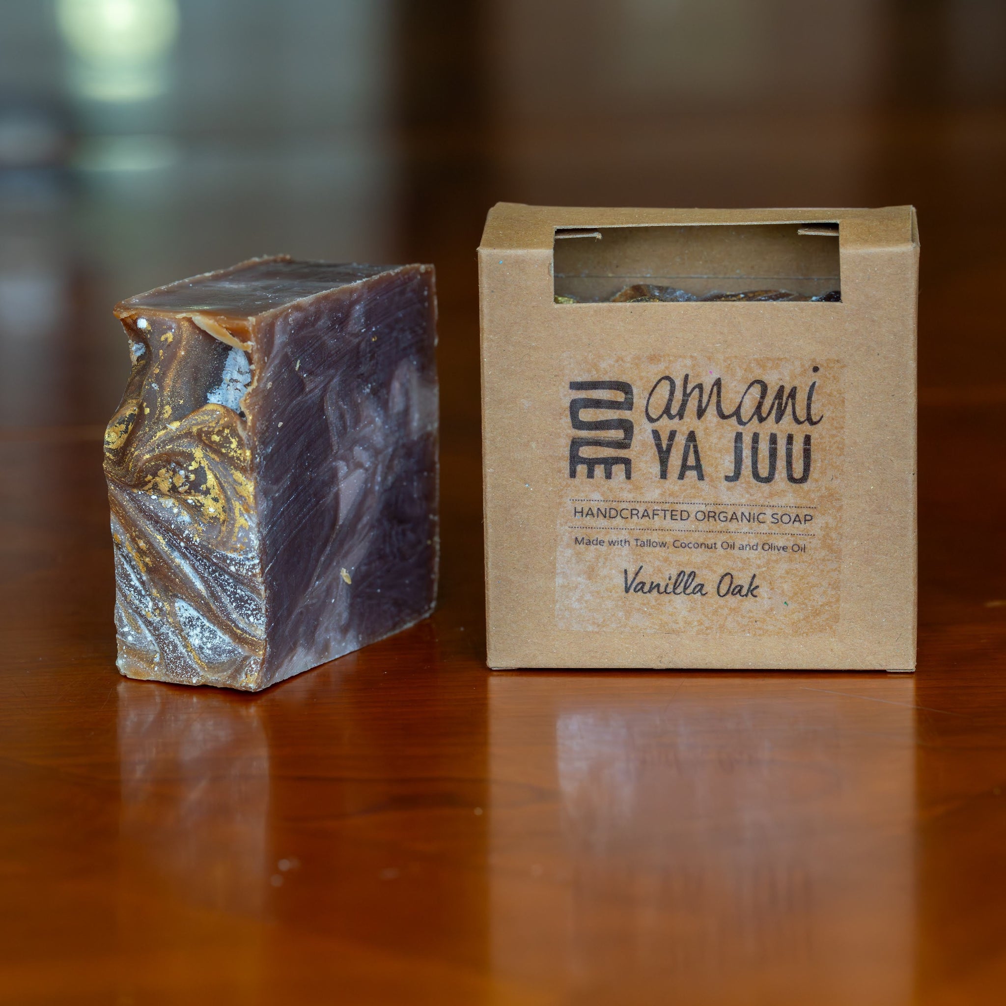 A rectangular bar of artisanal soap with a marbled purple and gold pattern sits next to its kraft paper packaging box on a polished wooden table. The box is printed with the brand name 'amani ya juu' and the scent 'Vanilla Oak'.