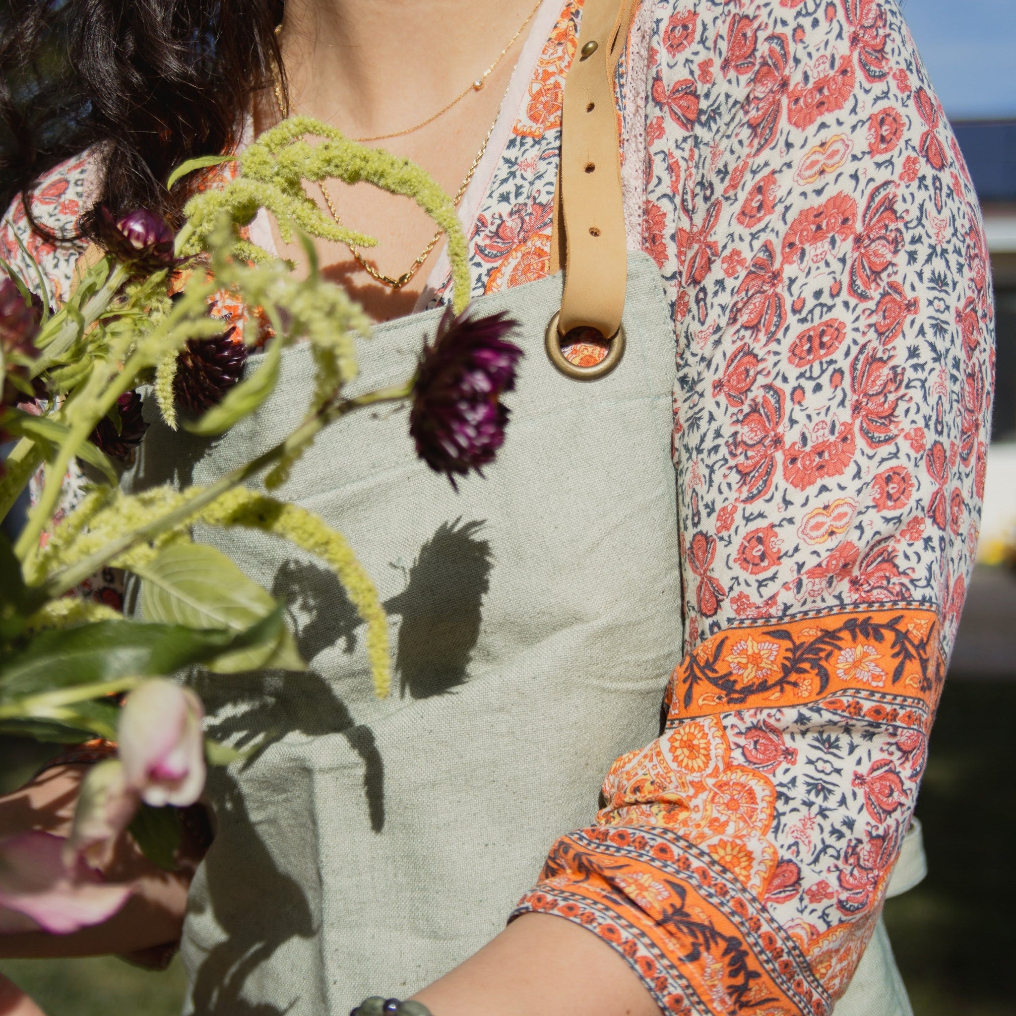 Person wearing a patterned shirt and green apron holding flowers with a blurred natural background