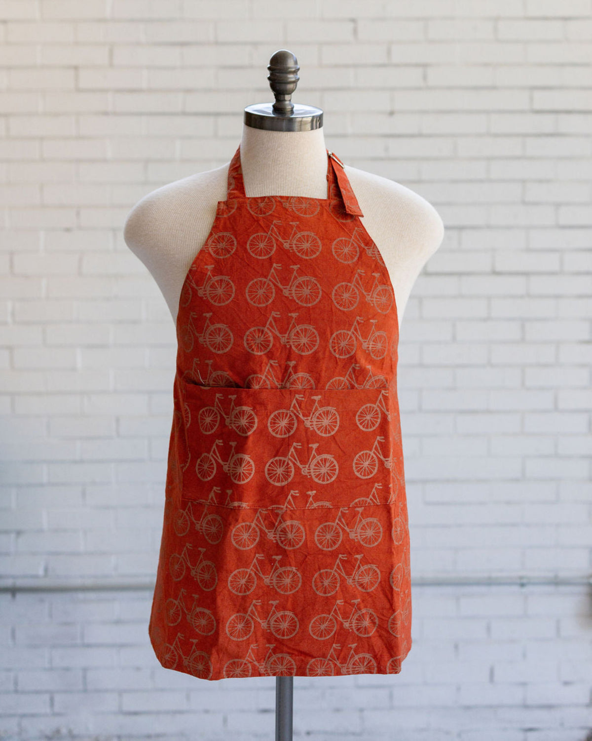 An orange halter-style apron with a repeating bicycle pattern, displayed on a cream-colored dress form mannequin against a white brick wall background.
