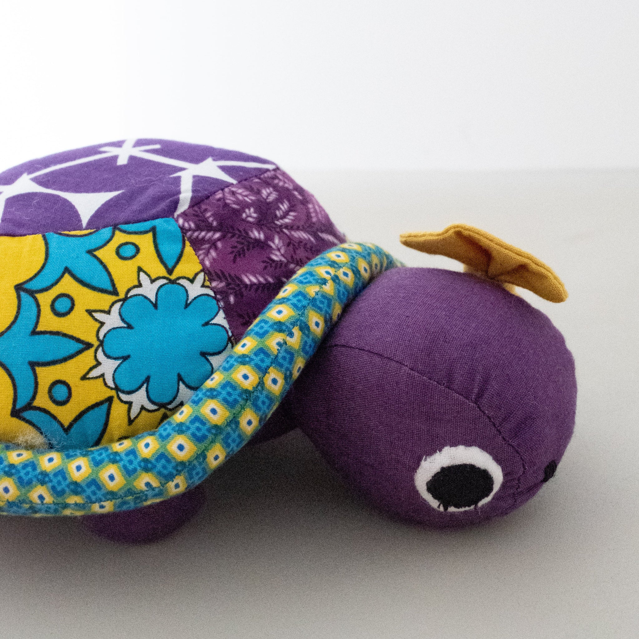 A handmade plush turtle toy made from colorful patterned fabrics, featuring a purple body and shell, a yellow and blue patterned border, and a small yellow accent on its head, set against a plain light gray background.