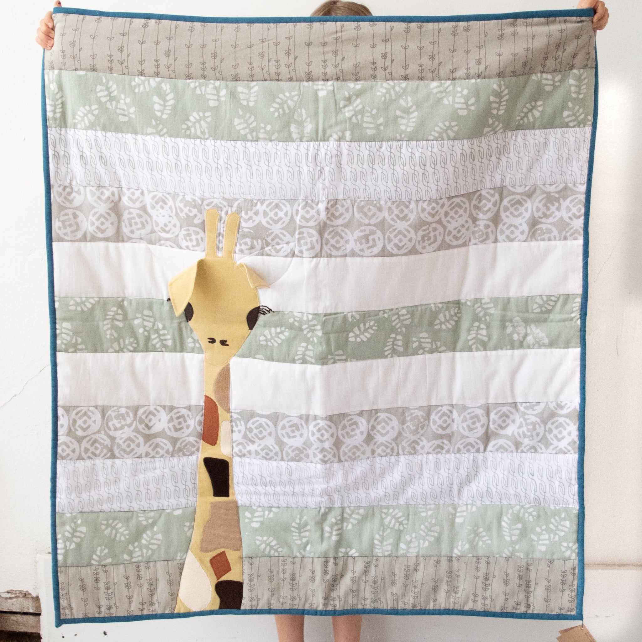 A person holding up a baby quilt featuring a giraffe design, with patterns of leaves and solid colors in shades of beige, brown, and white.