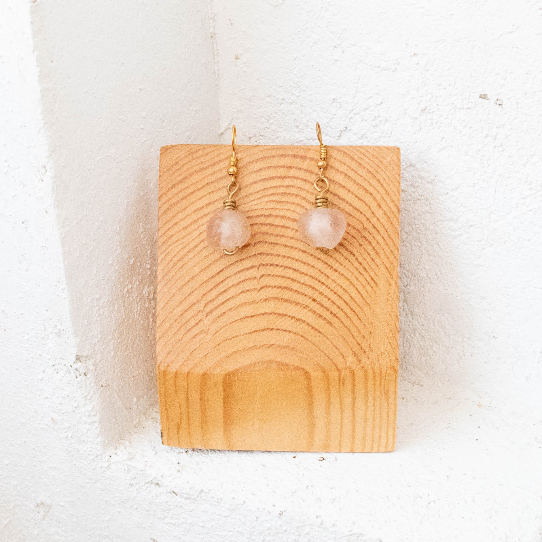 A pair of gold-tone dangle earrings with pale pink round beads, displayed on a small wooden stand against a white textured wall.