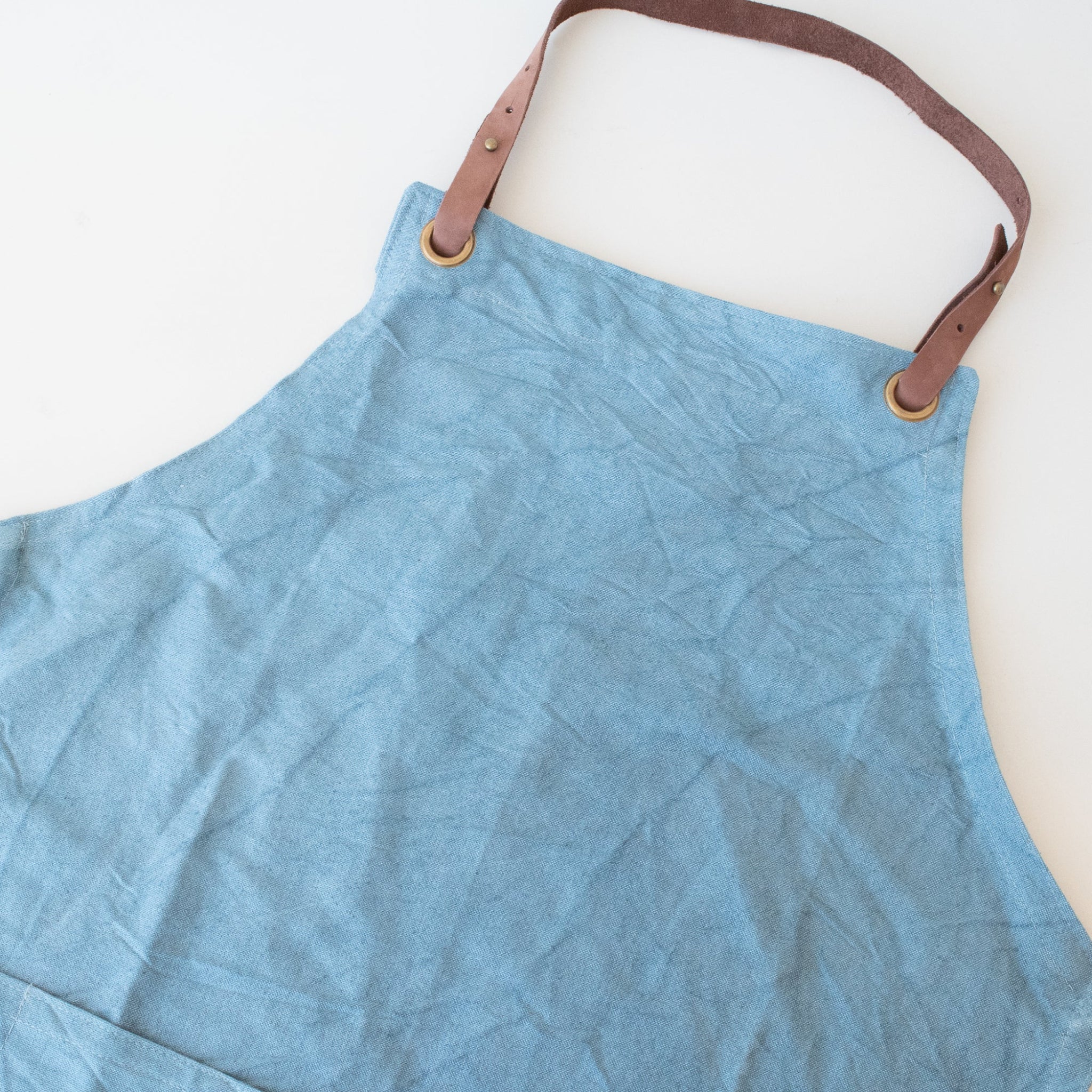 A light blue canvas apron with brown leather straps and brass grommets, laid flat on a white background.